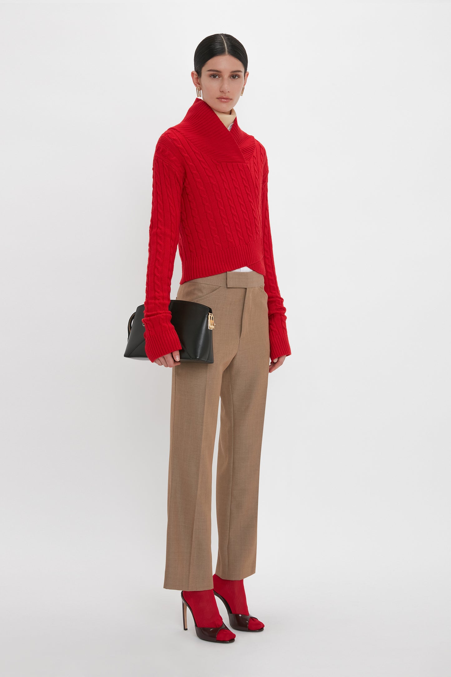 A person stands against a white background, wearing a red cable-knit sweater, Victoria Beckham Wide Cropped Flare Trouser In Tobacco, and red high heels, holding a black clutch bag.