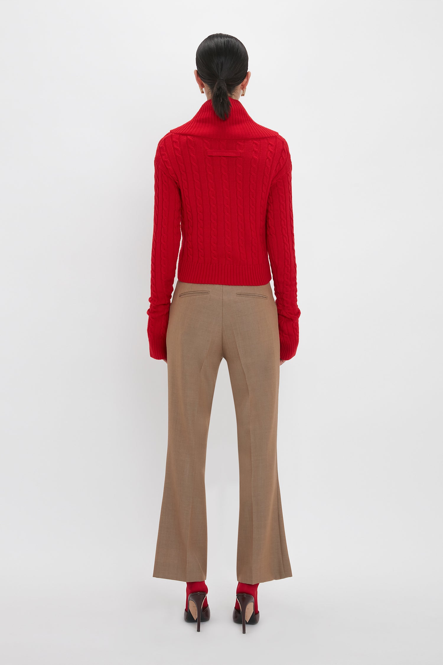 Individual standing against a plain backdrop, wearing a Victoria Beckham Wrap Detail Jumper In Red, tan trousers, and red high-heeled shoes, with hair tied back. Back view.