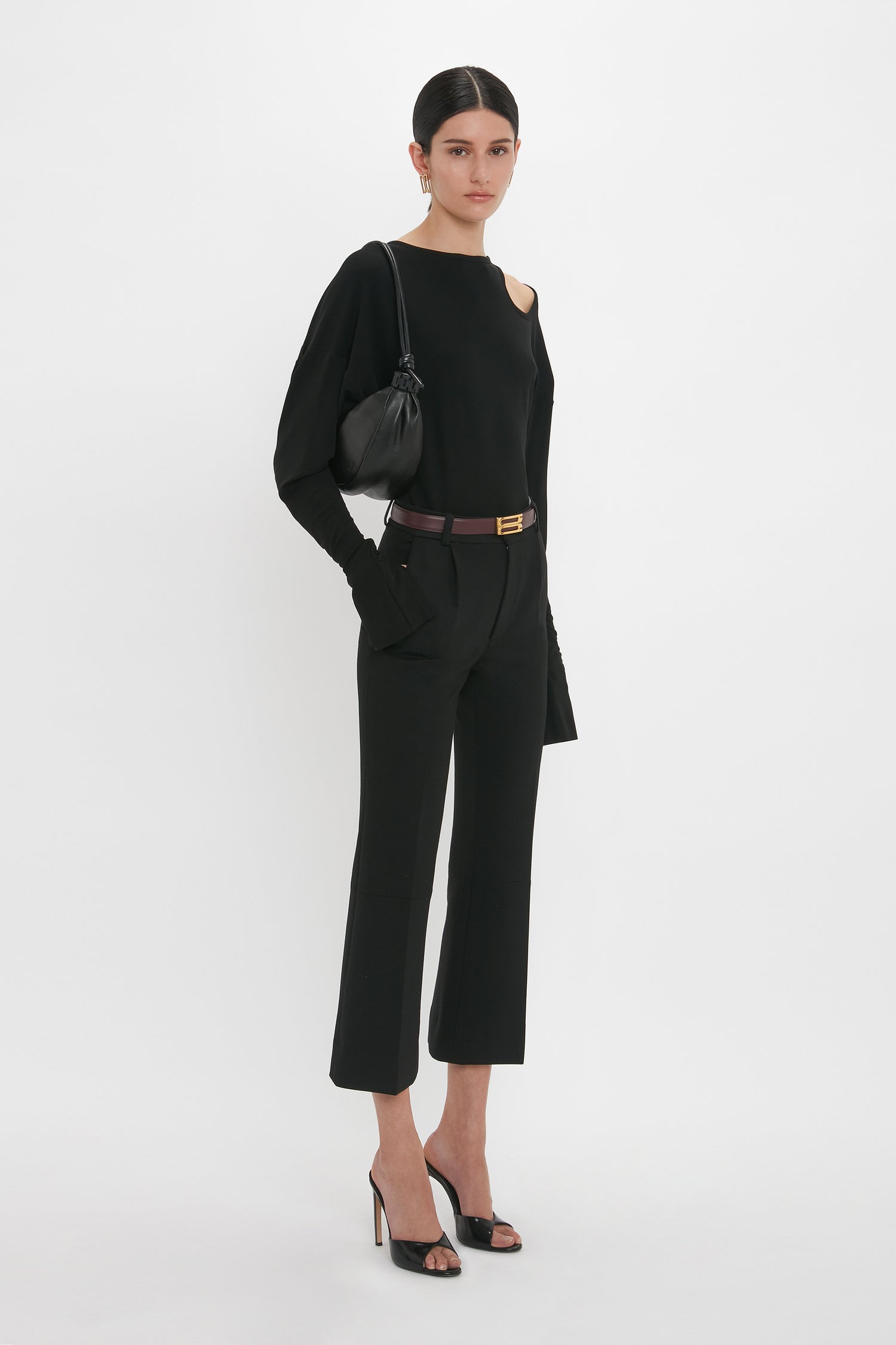 A person stands against a white background wearing a black Twist Detail Jersey Top In Black by Victoria Beckham with cut-out details. They hold a black bag over their shoulder and wear black high-heeled sandals, perfect for that wear-anywhere Black ensemble.