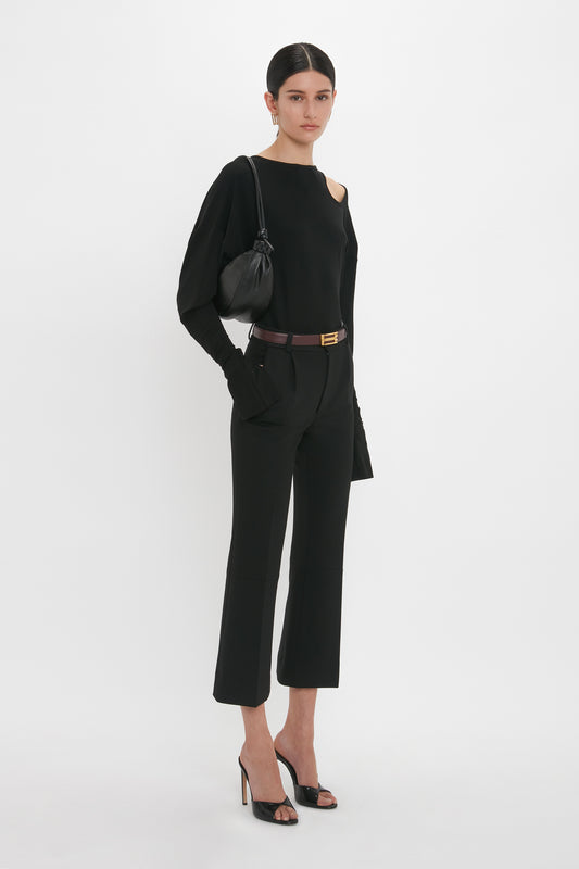 A woman stands against a white background wearing a black long-sleeve top, black cropped trousers, and Victoria Beckham Classic Mule In Black Calf Leather. She is holding a small black handbag over her shoulder.