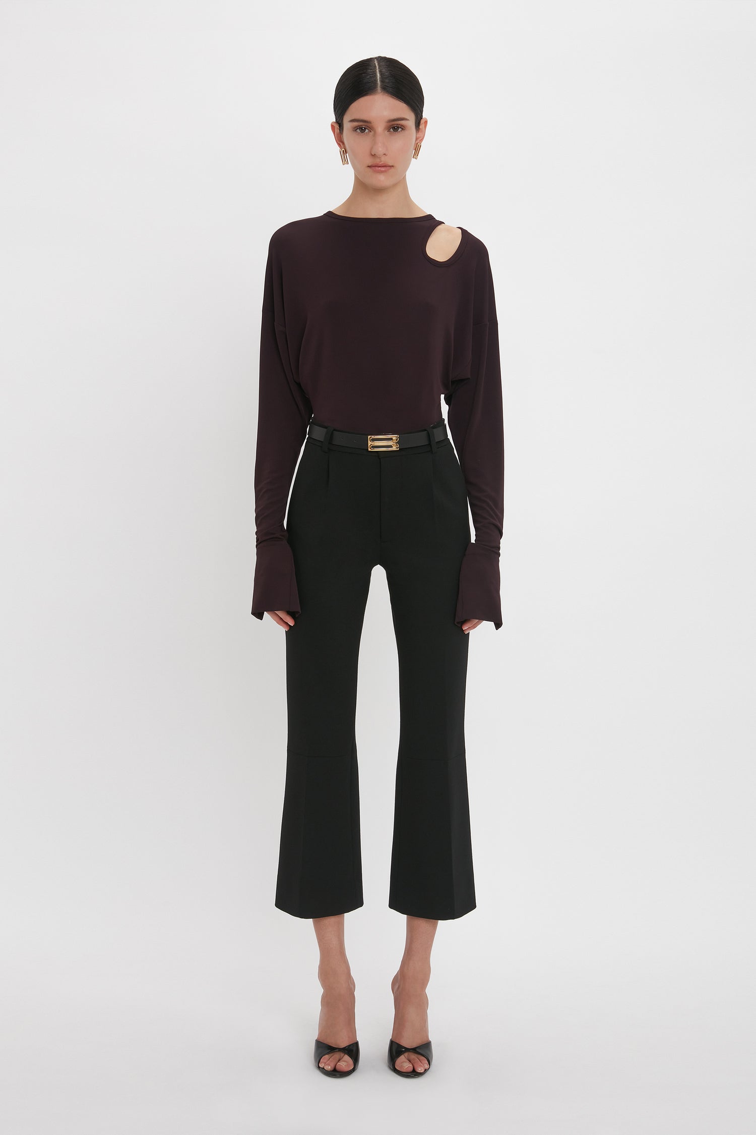 A person stands against a white background, embodying casual sophistication in a dark long-sleeve Twist Detail Jersey Top In Deep Mahogany by Victoria Beckham with a shoulder cut-out and black cropped pants. They are also wearing black open-toe heels.