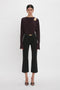 A person stands against a white background, embodying casual sophistication in a dark long-sleeve Twist Detail Jersey Top In Deep Mahogany by Victoria Beckham with a shoulder cut-out and black cropped pants. They are also wearing black open-toe heels.