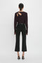 A person with dark hair in a ponytail is wearing a long-sleeved Victoria Beckham Twist Detail Jersey Top In Deep Mahogany with cutout details and black flared pants, exuding casual sophistication as they face away from the camera. They are also wearing black high-heeled shoes.