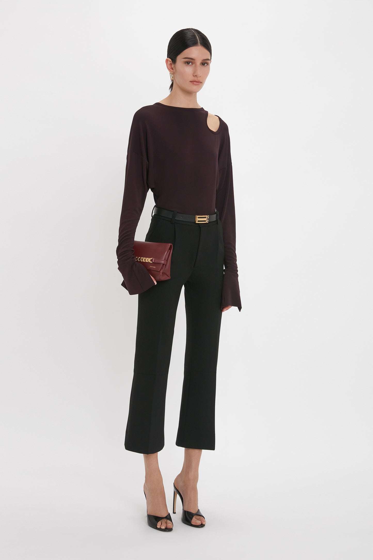 A woman stands against a plain background, epitomizing casual sophistication in a Twist Detail Jersey Top In Deep Mahogany by Victoria Beckham, black high-waisted pants, and black heeled sandals. She holds a small red clutch.