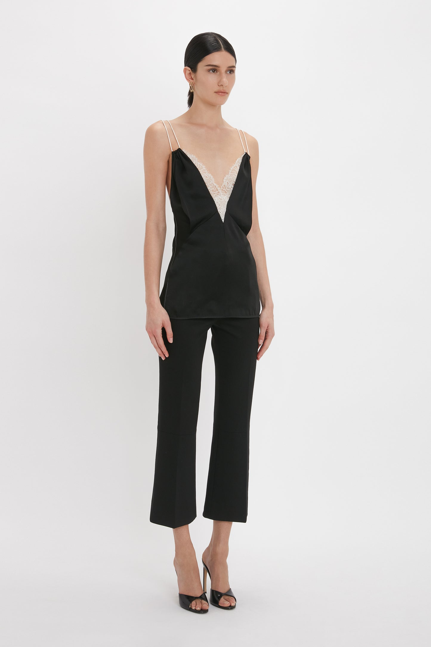 A person is standing against a plain background, wearing a Lace Detail Cami Top In Black by Victoria Beckham featuring a deep-V neckline and black cropped pants, paired with black high-heeled sandals.