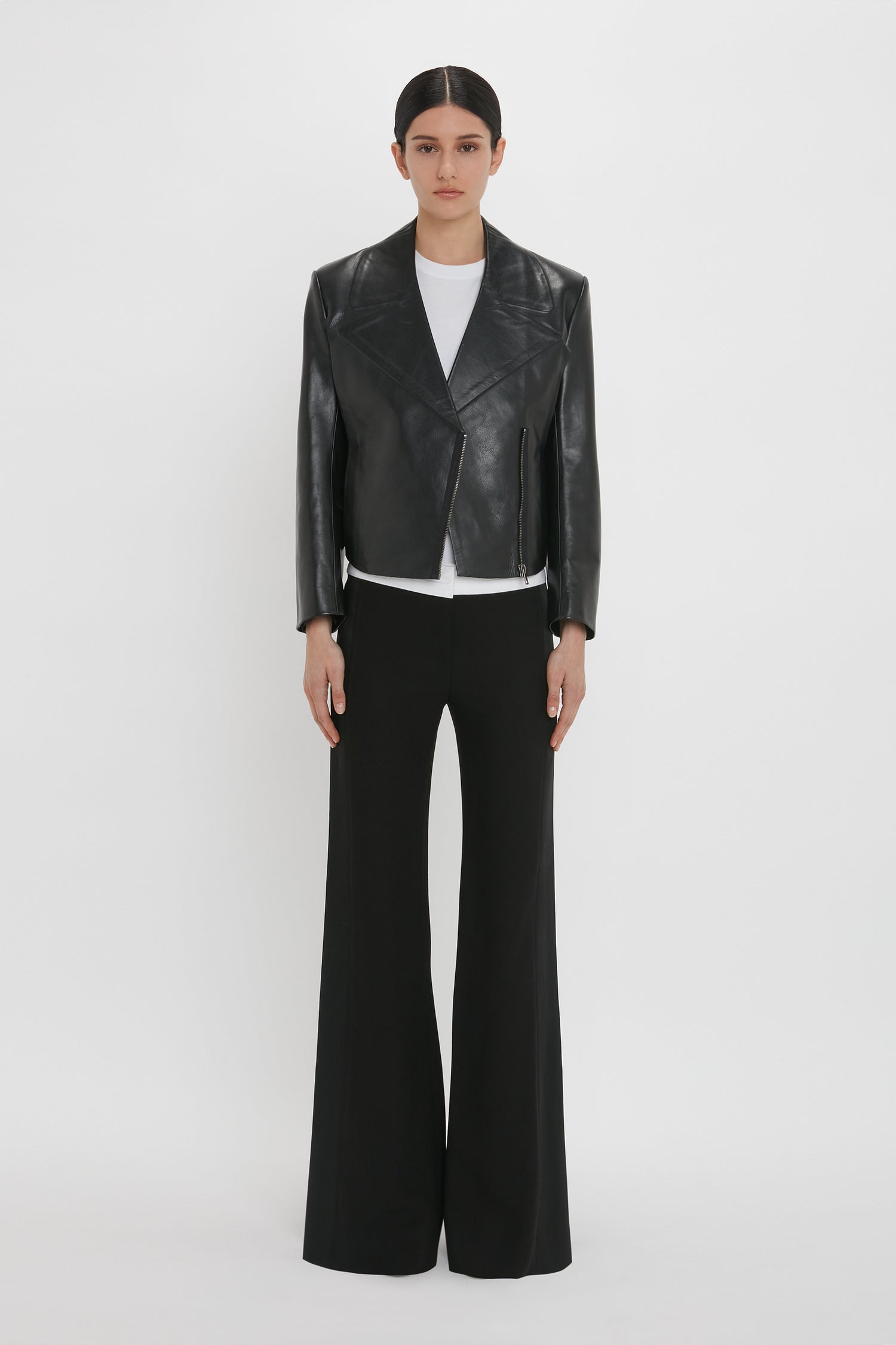 A person stands against a white background wearing a modern classic look: the Victoria Beckham Tailored Leather Biker Jacket In Black, a white shirt, and black wide-leg pants.