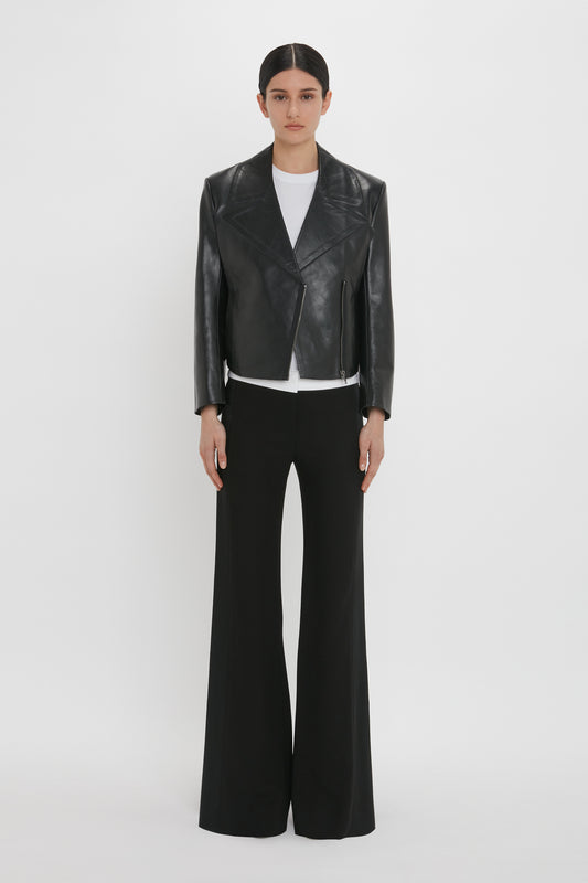 A person stands against a white background wearing a modern classic look: the Victoria Beckham Tailored Leather Biker Jacket In Black, a white shirt, and black wide-leg pants.