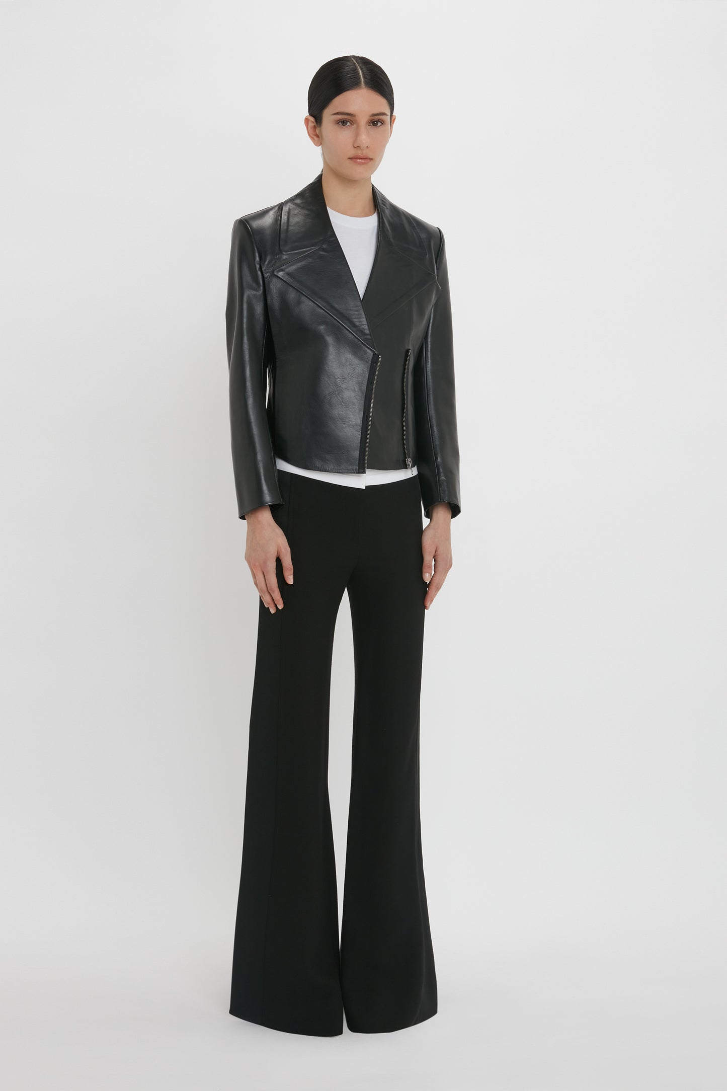 A person stands against a plain white background wearing a modern classic black leather jacket, white shirt, and black wide-leg pants. The Tailored Leather Biker Jacket In Black by Victoria Beckham and structured silhouette complete the stylish ensemble.