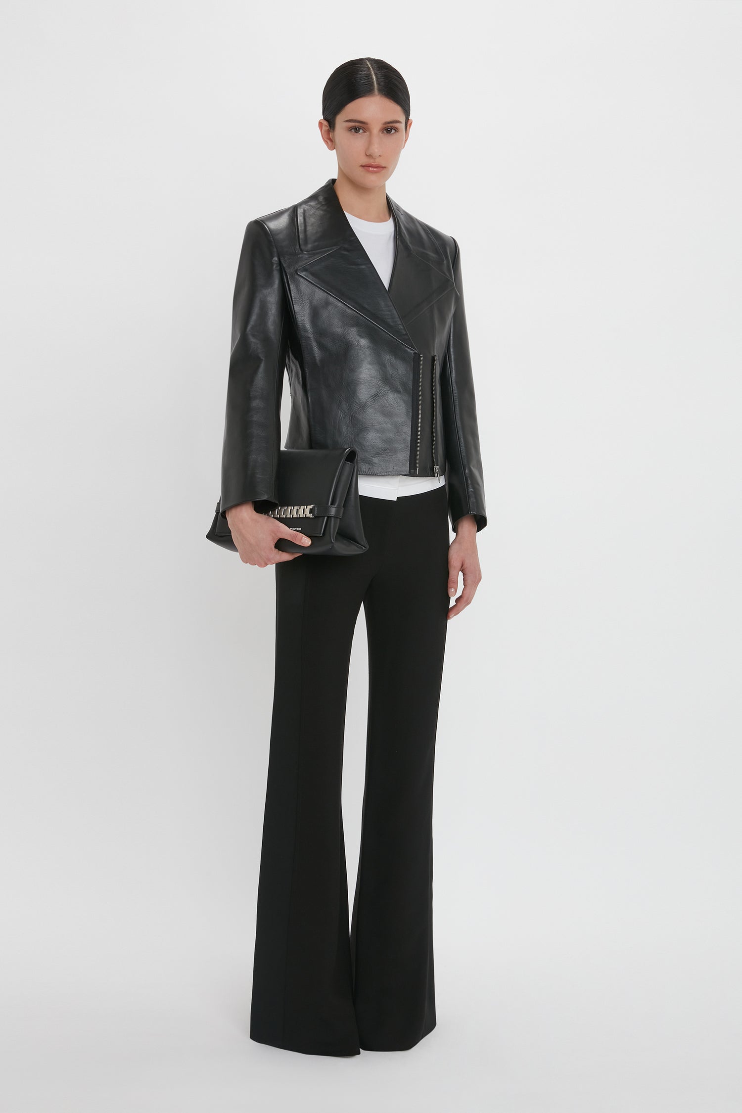 A person stands against a white background wearing a Victoria Beckham Tailored Leather Biker Jacket In Black, white shirt, and black pants, holding a black clutch bag.