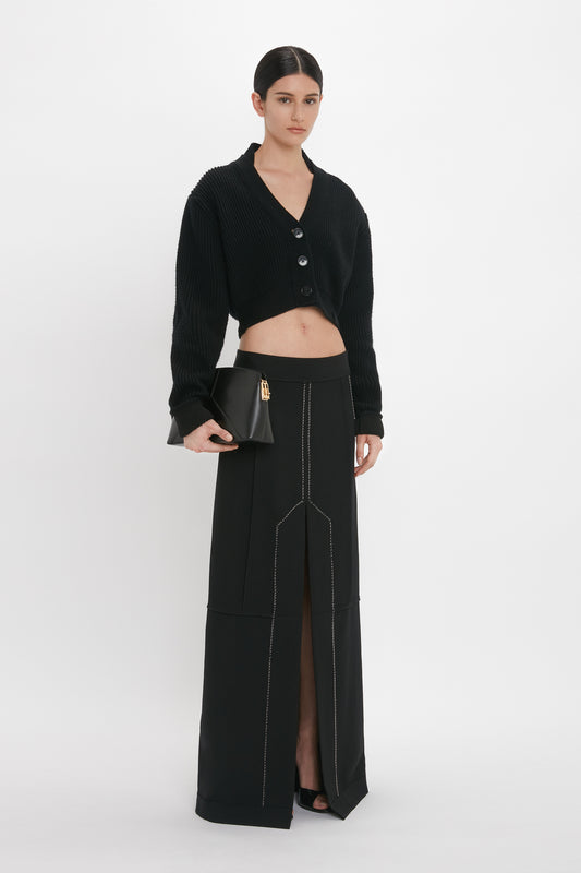 A woman stands in a Victoria Beckham Deconstructed Floor-Length Skirt In Black with a front slit, a black cropped cardigan with buttons, and holds a small black handbag. She has dark hair pulled back in a sleek style.