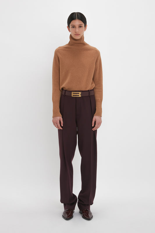 A person stands against a plain white background wearing a brown turtleneck sweater, Victoria Beckham Asymmetric Chino Trouser In Deep Mahogany in a narrow leg silhouette, and dark shoes. The individual has dark hair pulled back and is facing the camera.