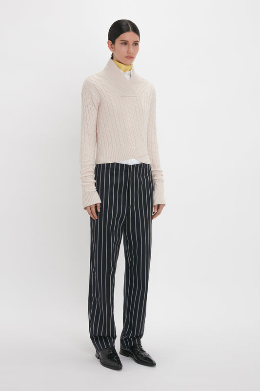 A person is standing against a plain white background, wearing a cream cable-knit sweater and black Victoria Beckham Tapered Leg Trouser In Midnight-White, paired with black shoes.