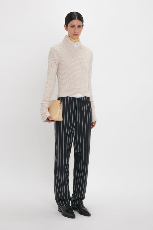 A person stands against a plain background, dressed in a cream knit sweater, Victoria Beckham's Tapered Leg Trouser In Midnight-White, and black shoes, holding a beige clutch bag.
