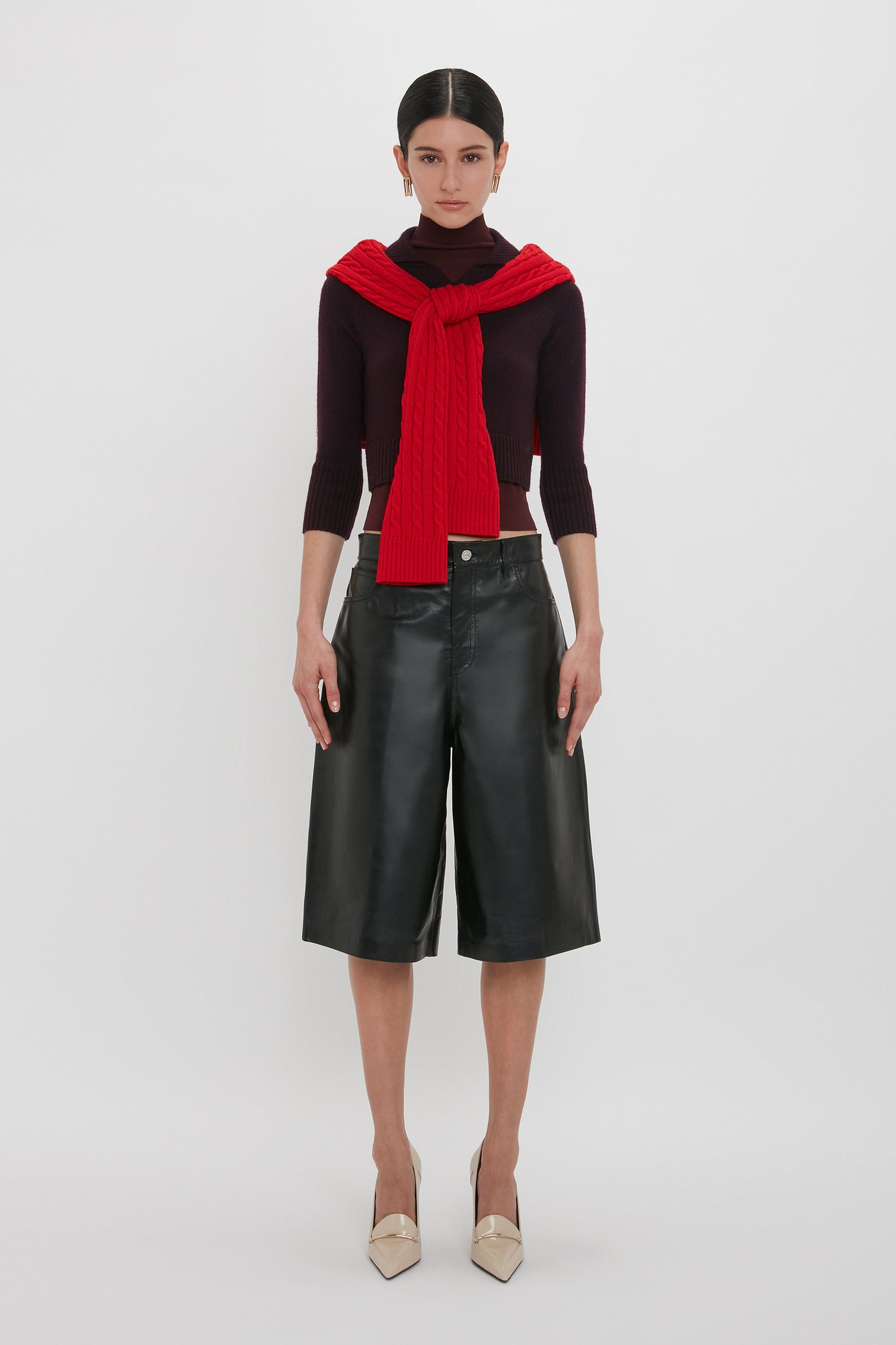 A person stands against a white background wearing a black turtleneck sweater, Leather Bermuda Short In Black by Victoria Beckham made of calf leather, and beige heels, with a red sweater draped over their shoulders.