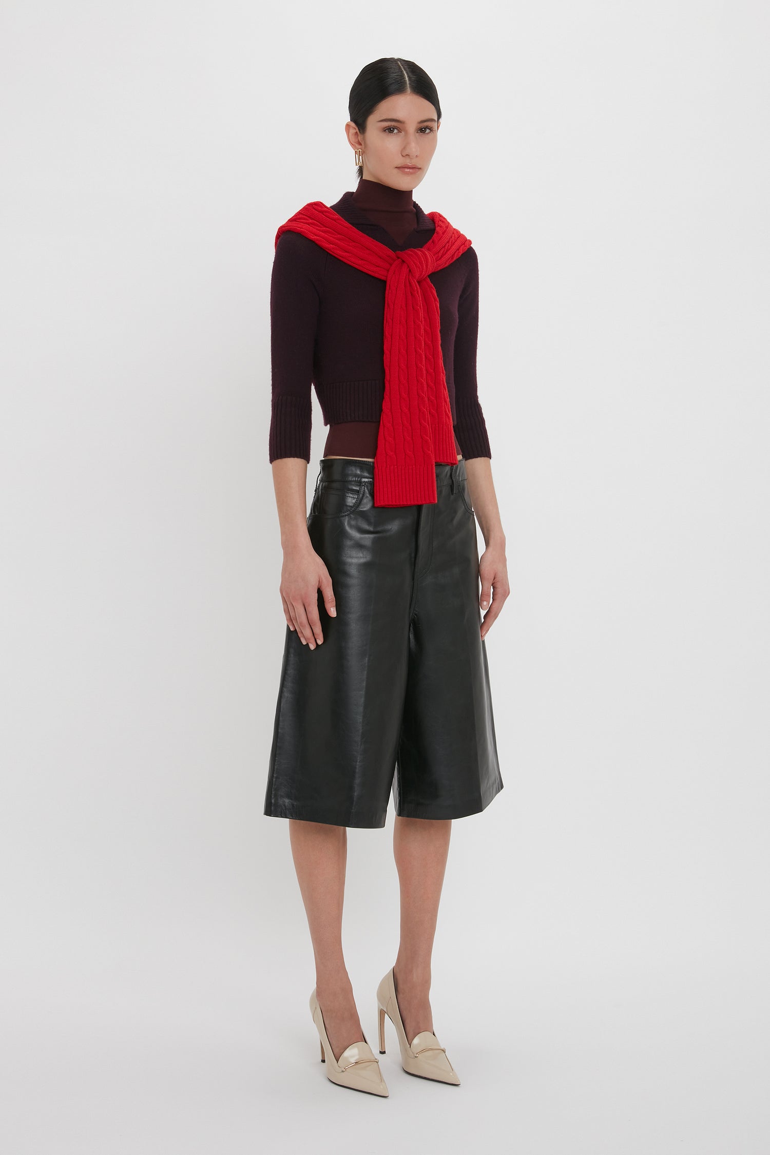 A person stands against a white background wearing a black turtleneck, Victoria Beckham's Leather Bermuda Short In Black that presents a flattering silhouette, a red sweater tied over the shoulders, and beige high heels.