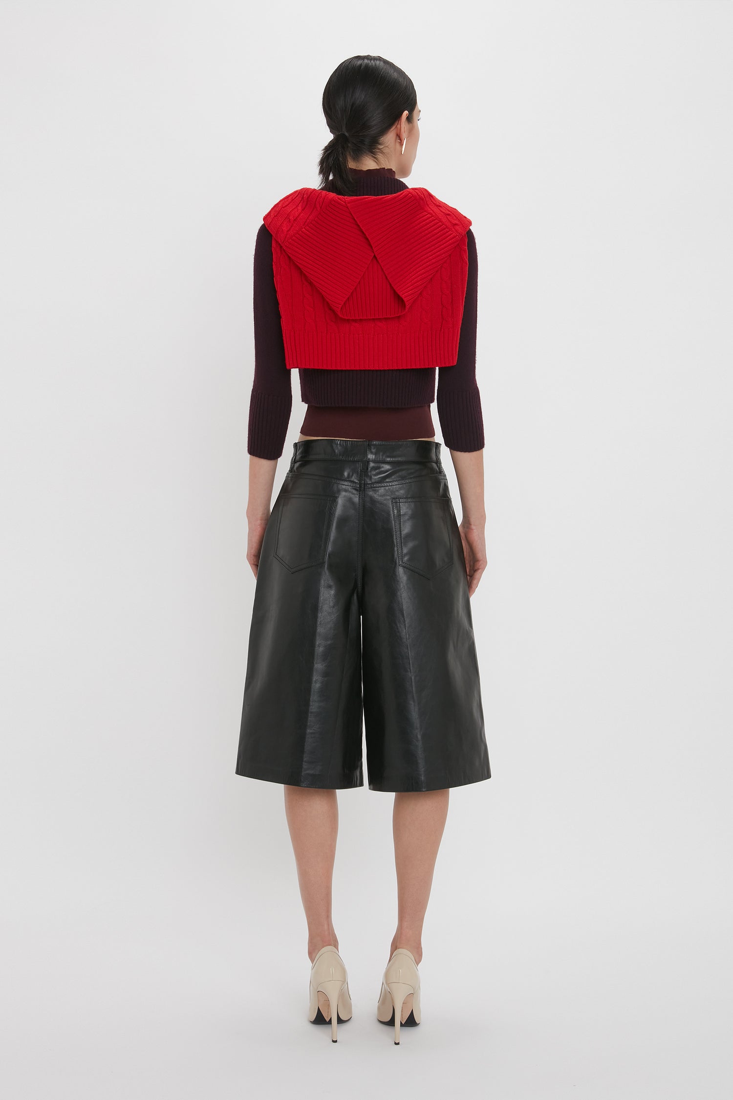 A woman is standing with her back to the camera, wearing a red sleeveless sweater over a long-sleeve top and beige high heels. Her Victoria Beckham Leather Bermuda Short In Black creates a flattering silhouette, paired perfectly with her tied-back hair.