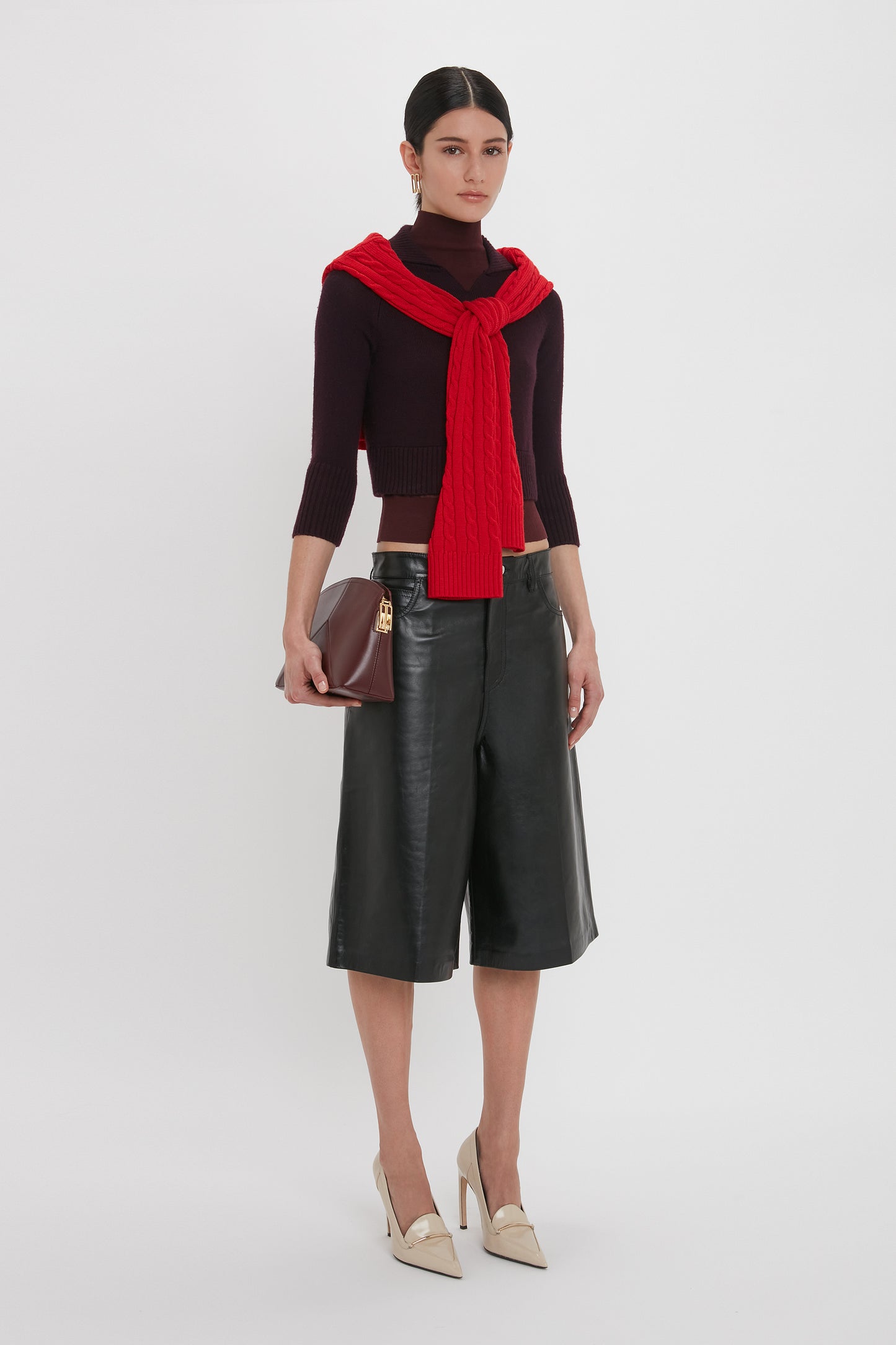 A woman wearing a black turtleneck, red sweater tied around her shoulders, Victoria Beckham Leather Bermuda Short In Black that create a flattering silhouette, beige heels, and holding a brown clutch stands against a white background.