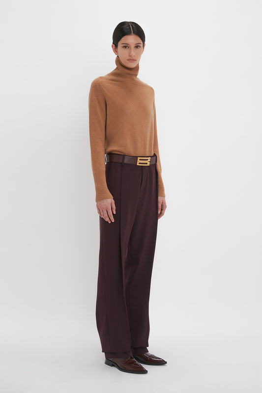 Person standing against a white background, wearing a brown turtleneck sweater, Victoria Beckham Asymmetric Chino Trouser In Deep Mahogany, and brown shoes.