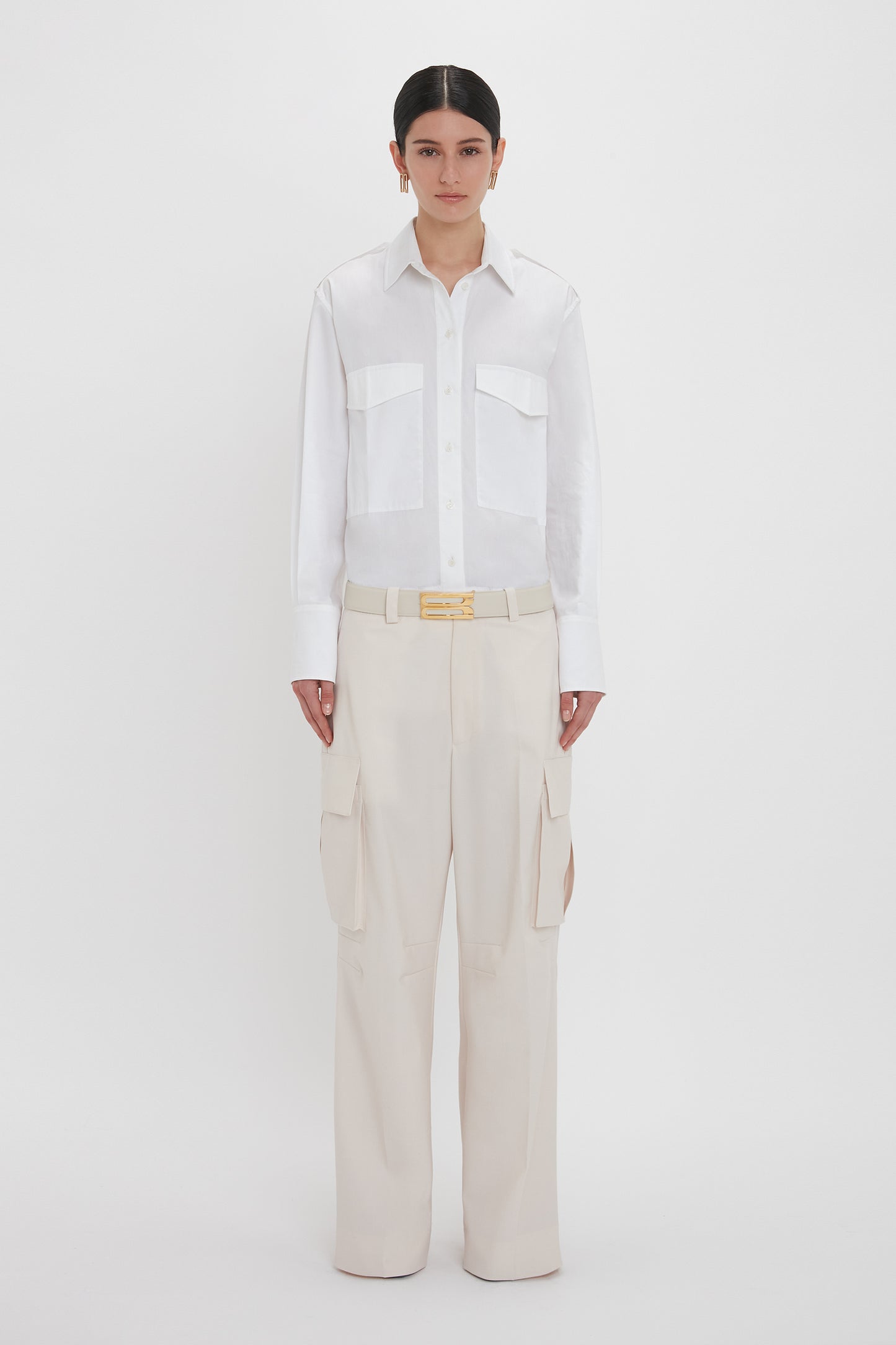 A person stands against a plain white background wearing an Oversized Pocket Shirt In White by Victoria Beckham made from organic cotton and beige cargo pants.