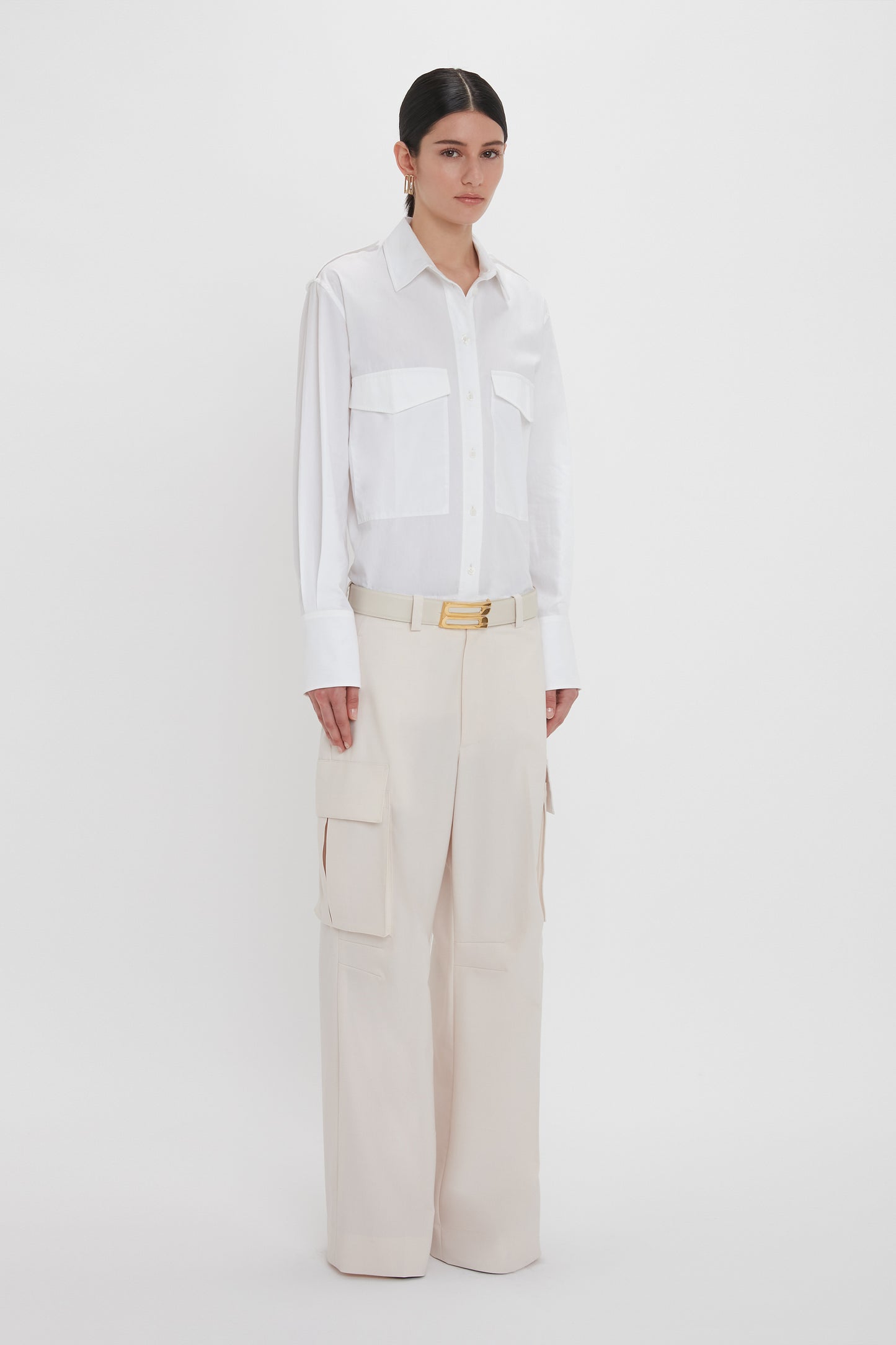 A person with dark hair wearing an Oversized Pocket Shirt in White by Victoria Beckham and beige high-waisted wide-leg cargo pants stands against a plain white background.