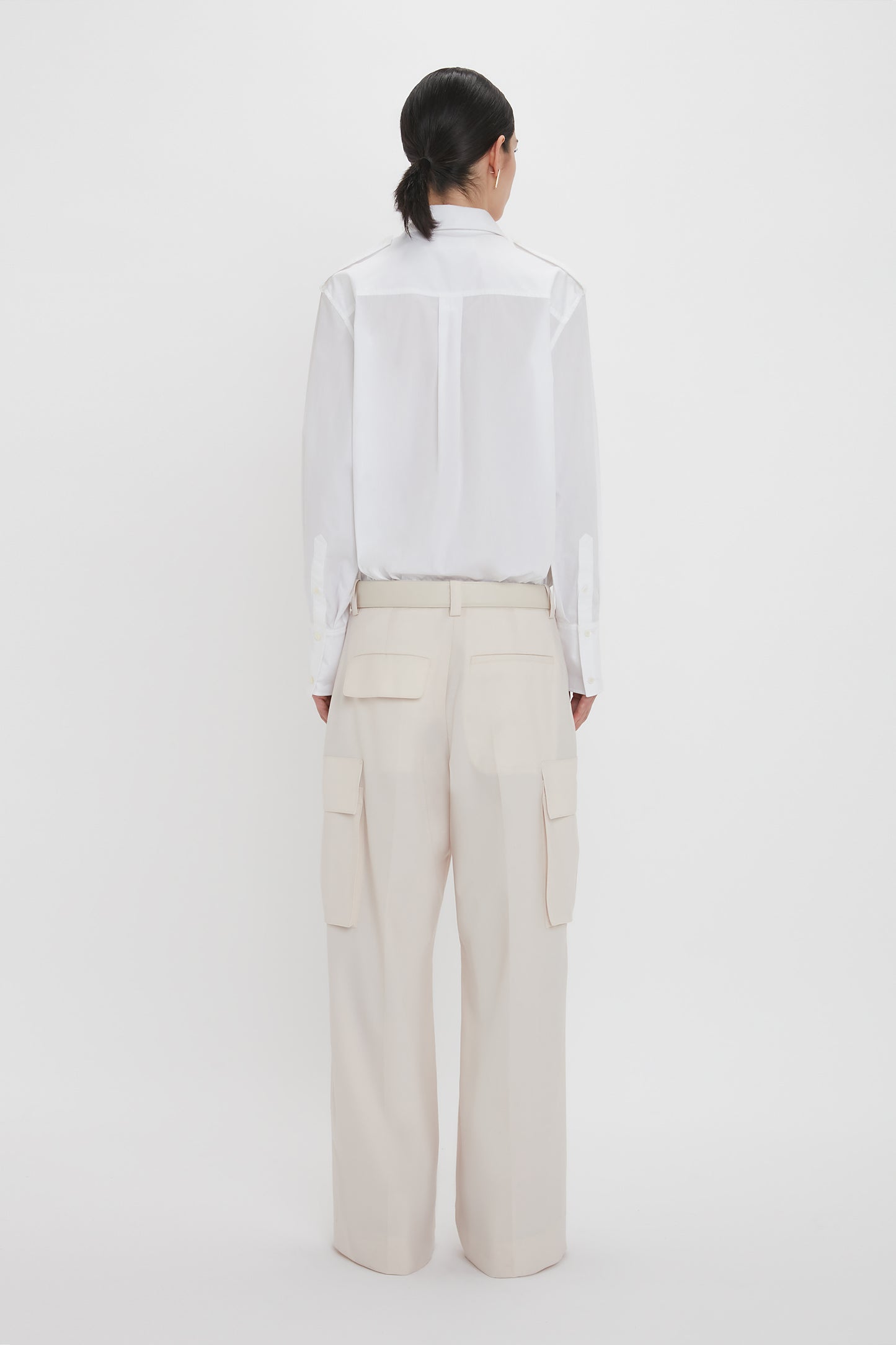 A person with dark hair, tied back, is standing with their back to the camera. They are wearing a Victoria Beckham Oversized Pocket Shirt In White and beige cargo pants with large pockets against a white background.
