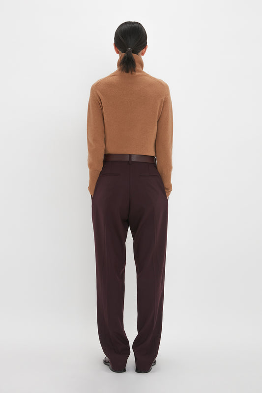 A person with dark hair tied back is seen from the back, wearing a brown cropped sweater and loose-fitting, high-waisted Victoria Beckham Asymmetric Chino Trouser In Deep Mahogany made from recycled wool, standing in front of a plain white background.