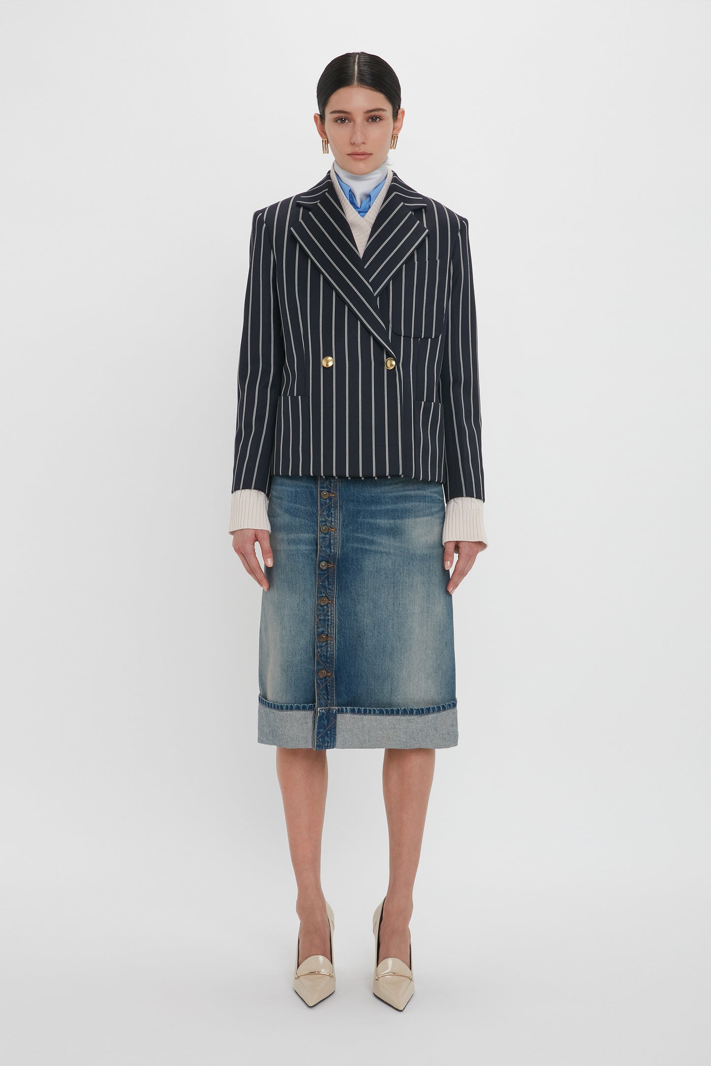 A person wearing a navy pinstripe double-breasted blazer, a light blue collared shirt with a scarf, a Victoria Beckham Placket Detail Denim Skirt In Heavy Vintage Indigo Wash, and beige pointed-toe heels stands against a plain white background.