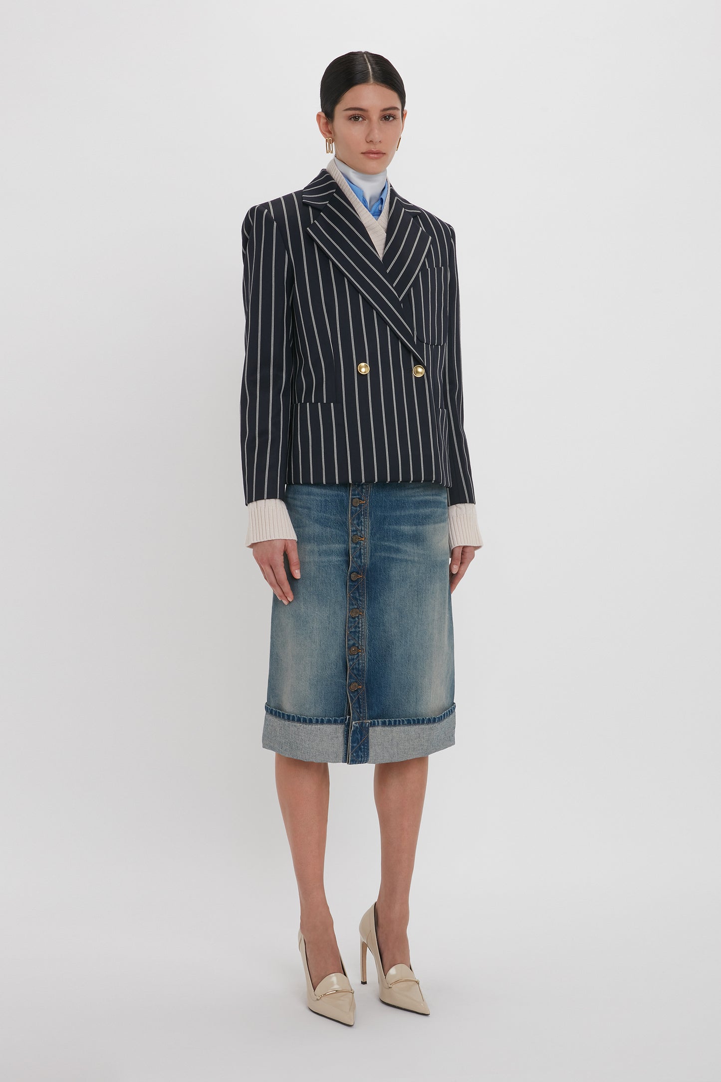 A woman stands against a plain background, wearing a navy pinstripe blazer, a white shirt, a blue necktie, a Placket Detail Denim Skirt In Heavy Vintage Indigo Wash by Victoria Beckham, and beige heels. Her hands are at her sides.