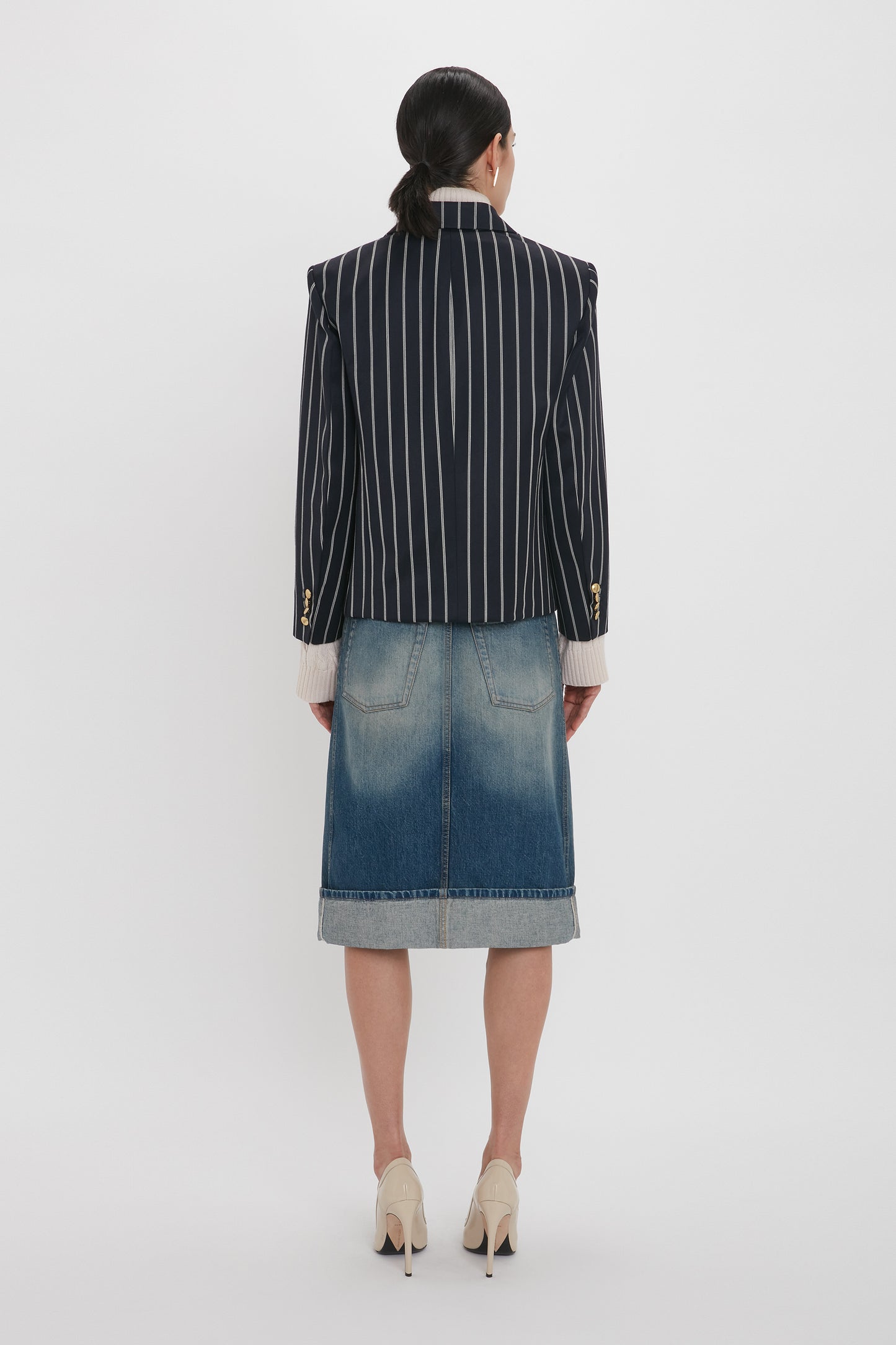 A person with long dark hair wearing a pinstriped blazer, a Placket Detail Denim Skirt In Heavy Vintage Indigo Wash by Victoria Beckham, and high heels stands facing a white background.