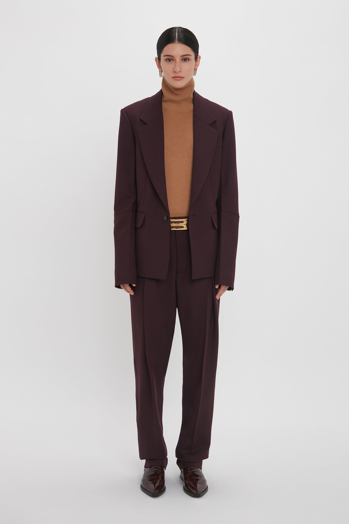 A person stands against a plain white background wearing the Sleeve Detail Patch Pocket Jacket In Deep Mahogany by Victoria Beckham, along with a deep mahogany turtleneck and matching brown dress shoes. They have straight dark hair tied back and wear minimal jewelry.