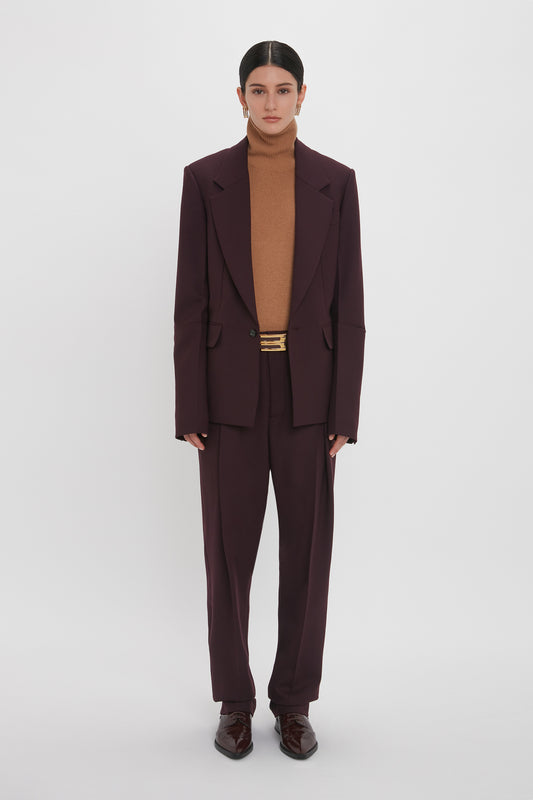 A person stands against a plain white background wearing the Sleeve Detail Patch Pocket Jacket In Deep Mahogany by Victoria Beckham, along with a deep mahogany turtleneck and matching brown dress shoes. They have straight dark hair tied back and wear minimal jewelry.