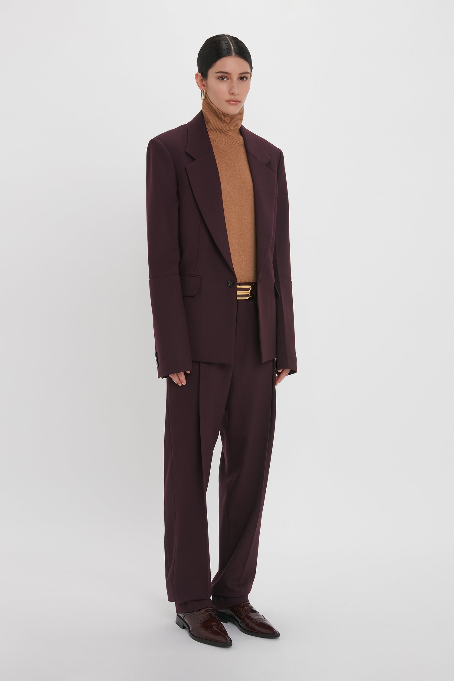 Person standing against a white background, wearing a dark burgundy Sleeve Detail Patch Pocket Jacket In Deep Mahogany by Victoria Beckham featuring a single-breasted blazer with a brown turtleneck sweater underneath and dark brown shoes.