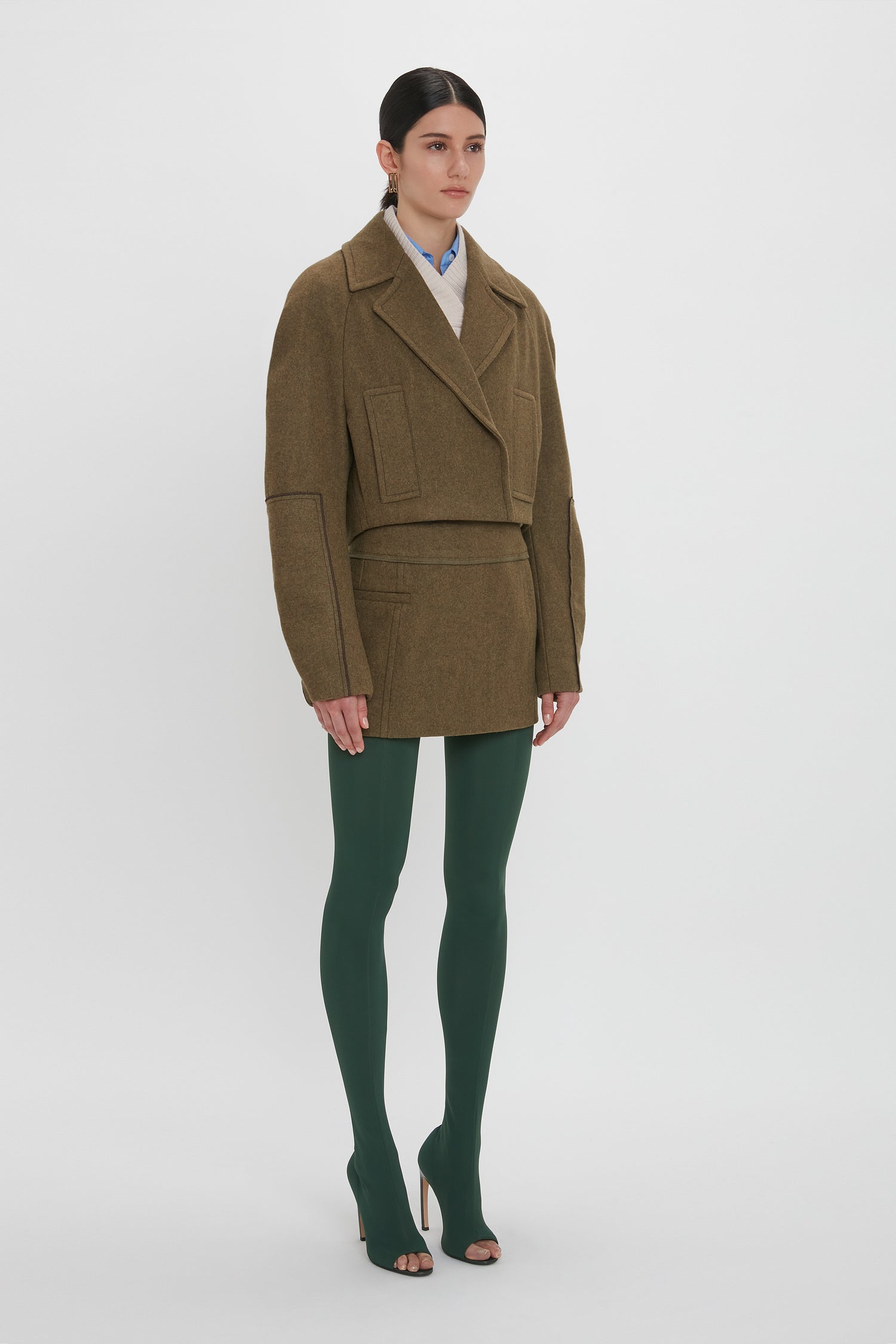 A person is wearing a Cropped Pea Coat In Khaki by Victoria Beckham, a matching mini skirt, and green tights made of Merino Wool. They complete their look with open-toe high heels and are standing against a plain white background.