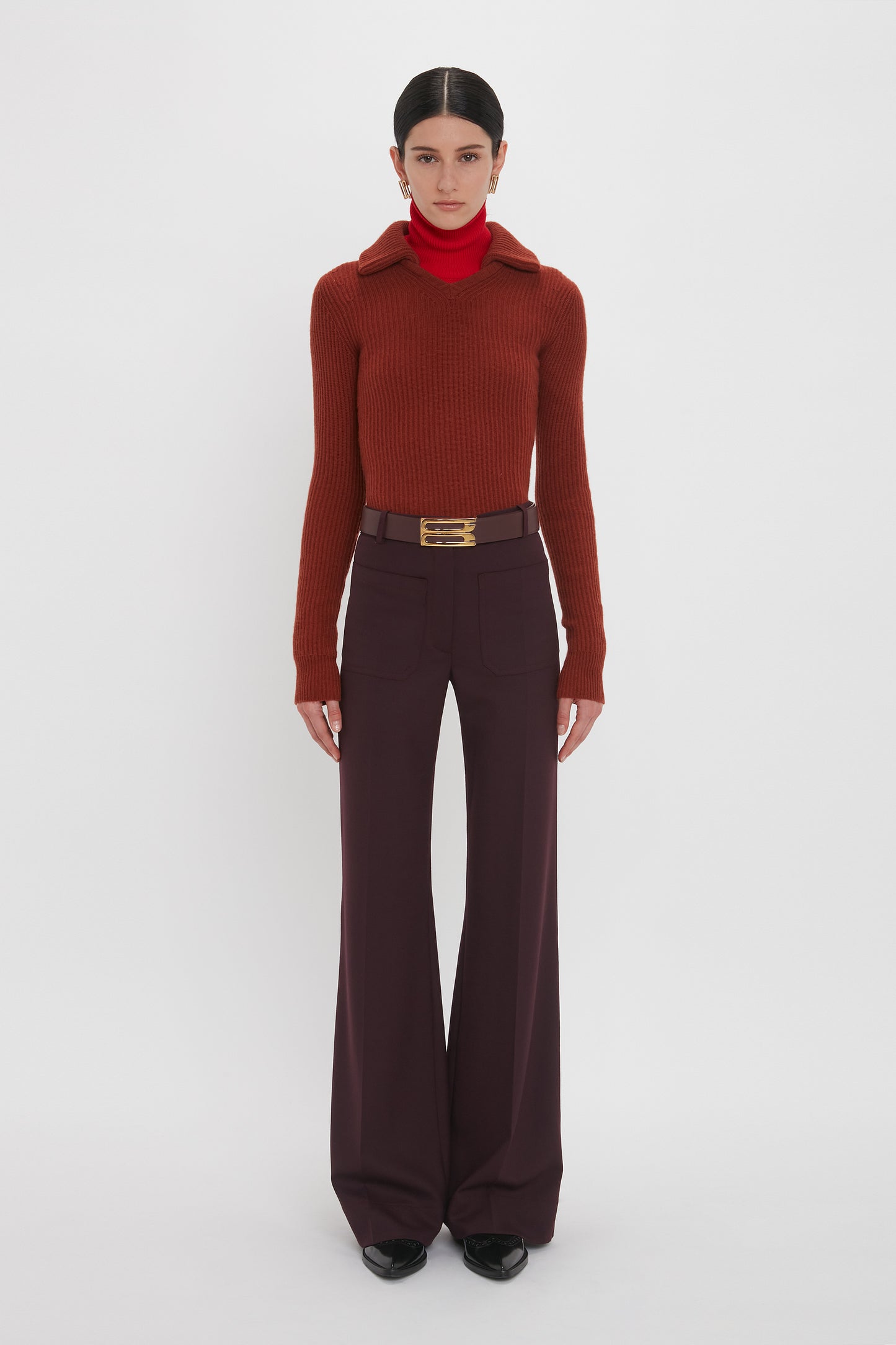 A person stands facing the camera, clad in a Double Collared Jumper In Russet by Victoria Beckham over a red turtleneck and dark wide-leg trousers with a belt. The background is plain white.