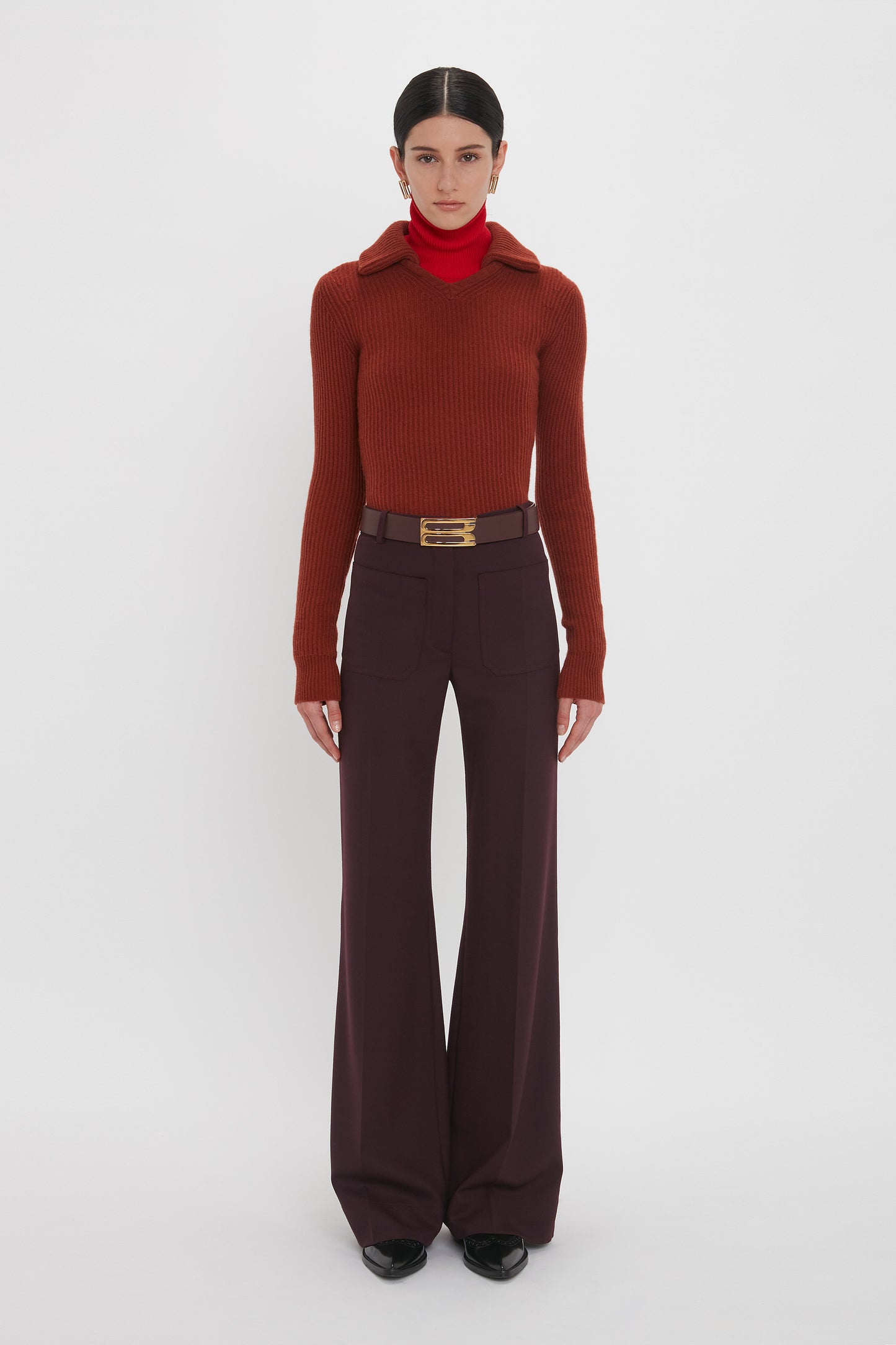 A person wearing a red turtleneck under a rust-colored recycled wool sweater paired with high-waisted deep mahogany Alina Trousers by Victoria Beckham and a belt. The person stands facing forward against a plain white background.
