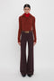 A person wearing a red turtleneck under a rust-colored recycled wool sweater paired with high-waisted deep mahogany Alina Trousers by Victoria Beckham and a belt. The person stands facing forward against a plain white background.