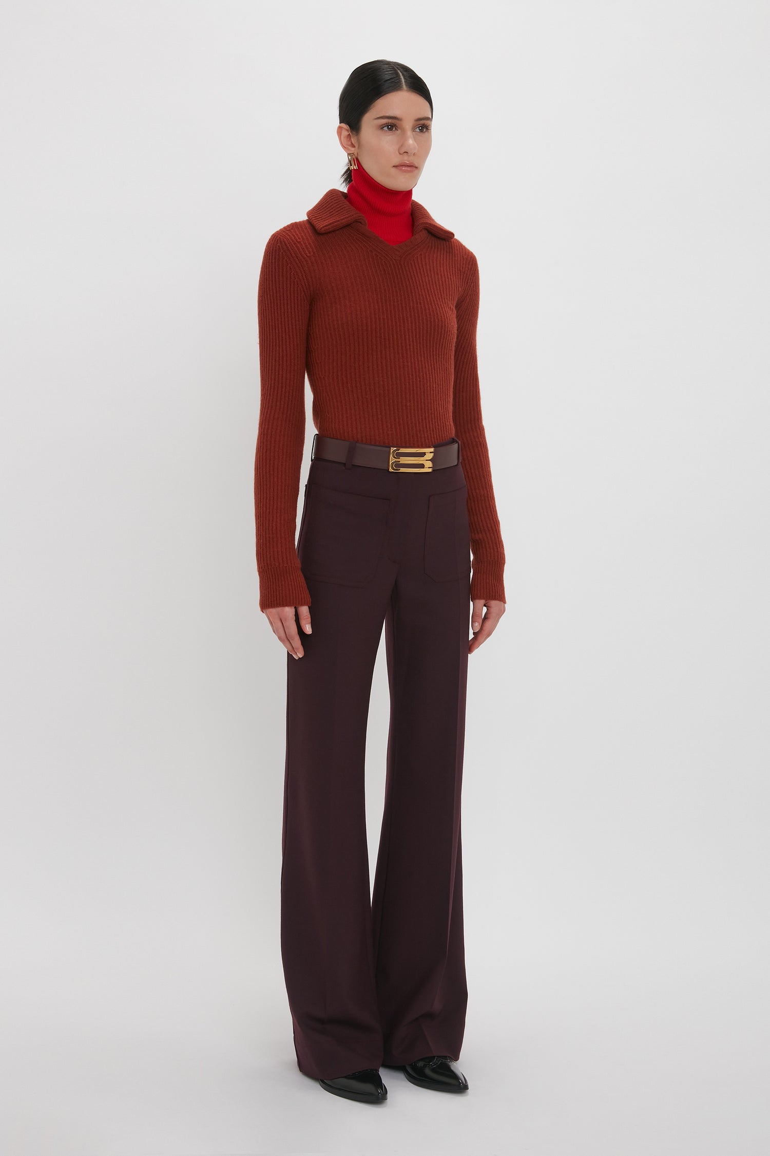 A person stands against a plain white background wearing a Double Collared Jumper In Russet by Victoria Beckham, maroon wide-legged trousers, a red turtleneck, black shoes, and a brown belt with a gold buckle.