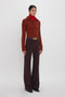 A woman stands against a plain white background, wearing a red ribbed sweater, a red turtleneck, and high-waisted, wide-leg Alina Trouser In Deep Mahogany made from recycled wool with a brown belt. This Victoria Beckham-inspired look effortlessly combines style and sustainability.