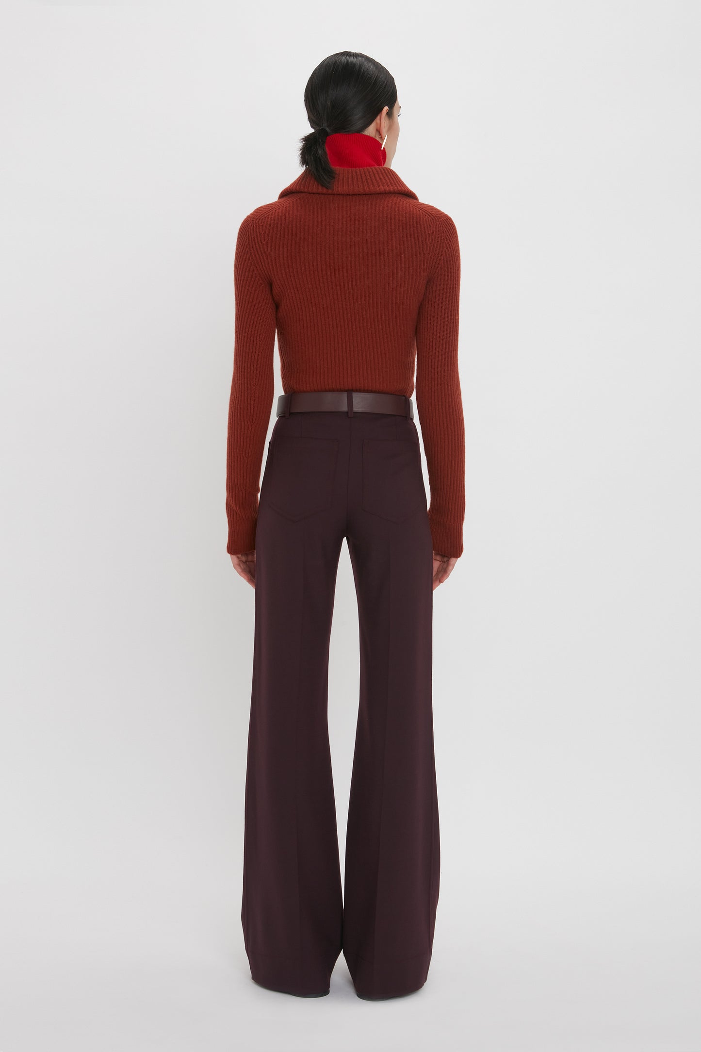 Person with dark hair in a ponytail, wearing a red turtleneck sweater and high-waisted, wide-leg Victoria Beckham Alina Trouser In Deep Mahogany pants made from recycled wool, viewed from the back against a plain white background.
