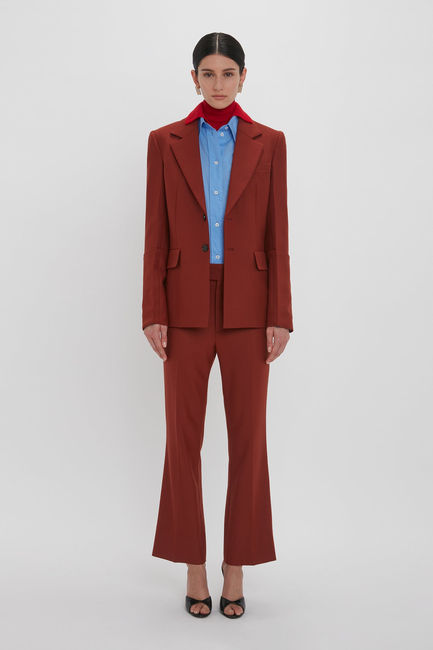 Person standing against a neutral background, dressed in a rust-colored suit with 1970s-inspired **Wide Cropped Flare Trouser In Russet by Victoria Beckham**, a blue shirt, a red ribbon around the neck, and black open-toe heels.