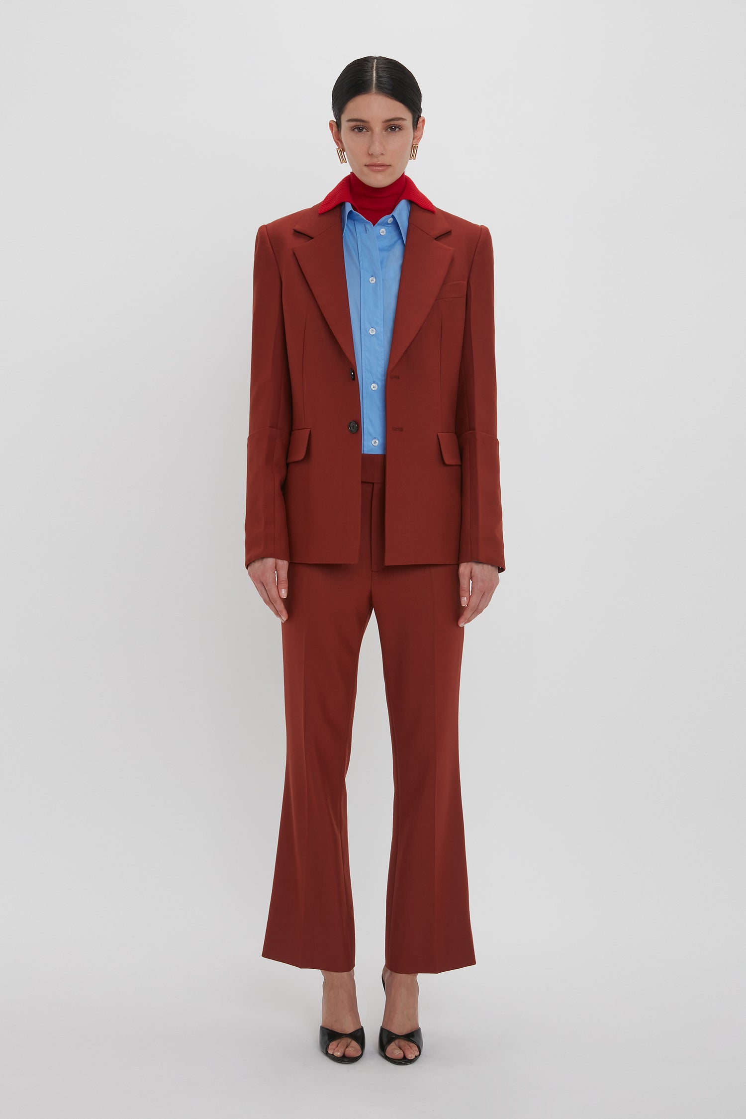 A person is standing against a plain background, wearing a rust-colored suit featuring contemporary detailing with a blue shirt and red scarf. They are also wearing the Sleeve Detail Patch Pocket Jacket In Russet by Victoria Beckham.