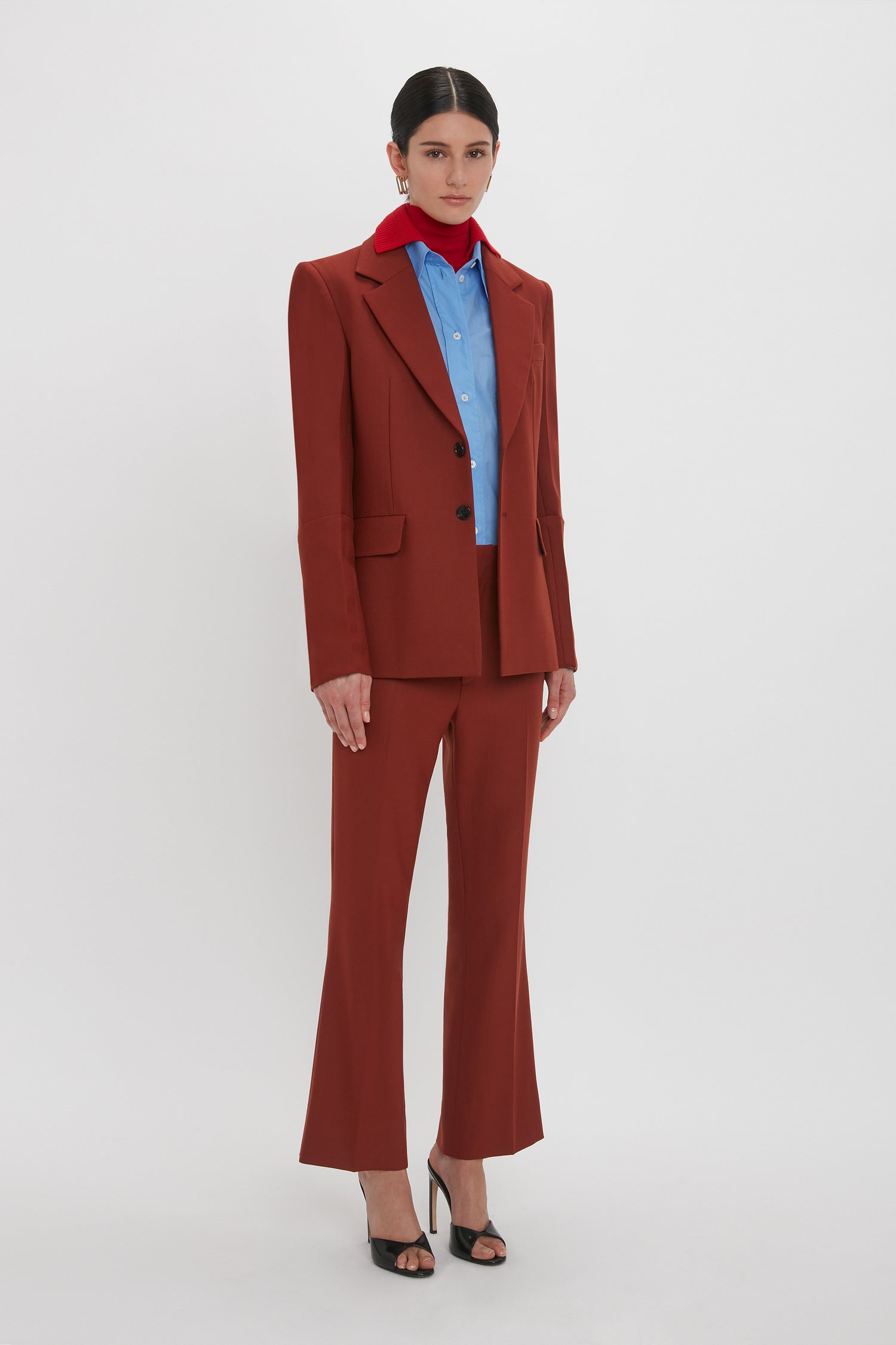 Person standing against a white background, wearing a Victoria Beckham Sleeve Detail Patch Pocket Jacket In Russet over a blue shirt and red scarf, paired with black high-heeled shoes. The outfit merges contemporary detailing seamlessly.
