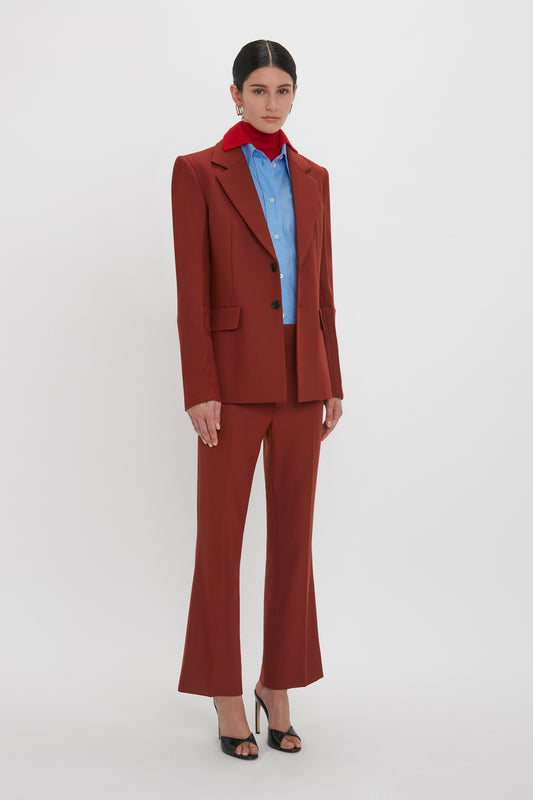 Person standing against a white background, wearing a Victoria Beckham Sleeve Detail Patch Pocket Jacket In Russet over a blue shirt and red scarf, paired with black high-heeled shoes. The outfit merges contemporary detailing seamlessly.