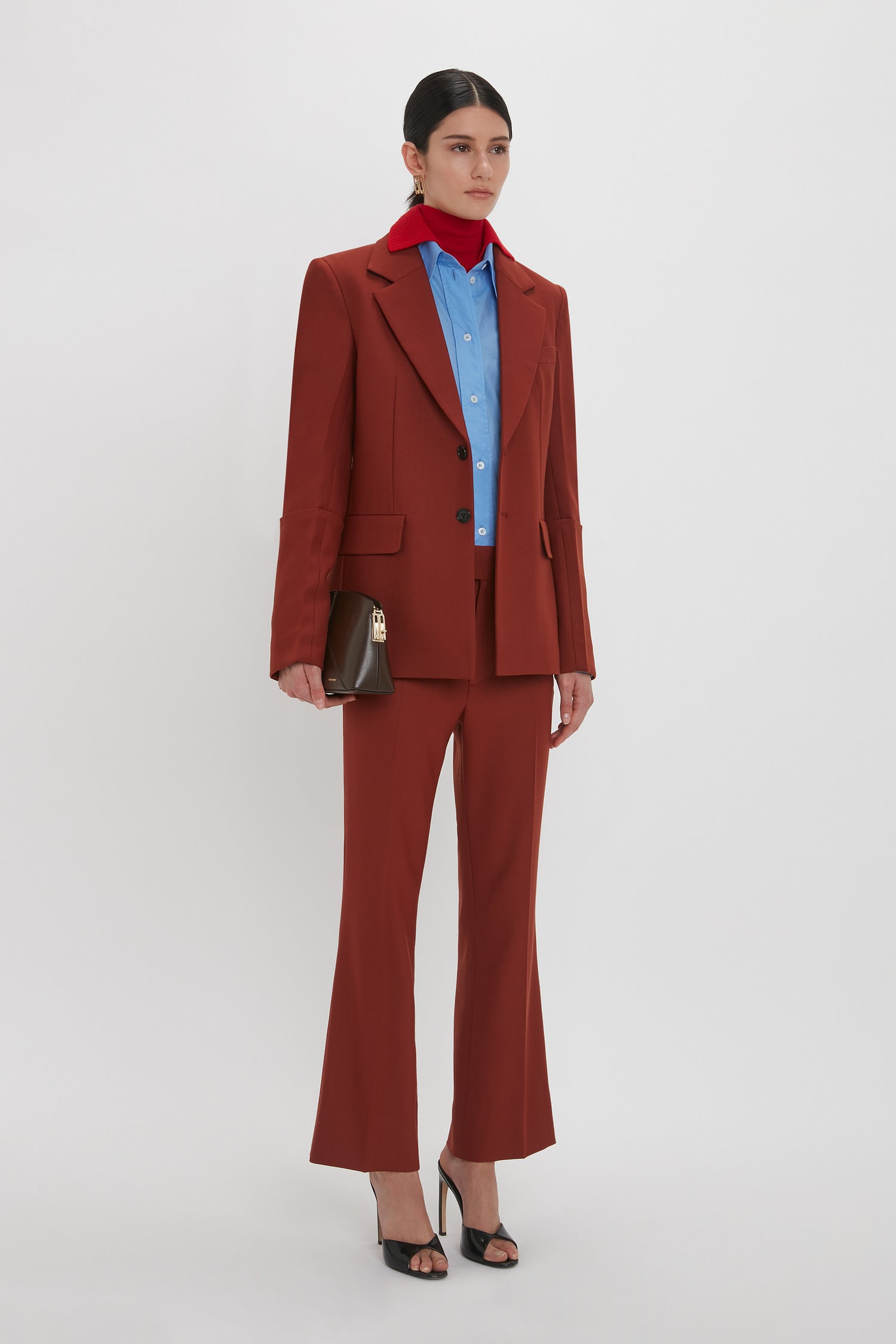 Person wearing a dark red Wide Cropped Flare Trouser In Russet by Victoria Beckham with 1970s-inspired trousers and a blue shirt, holding a brown clutch, and wearing black open-toe heels.