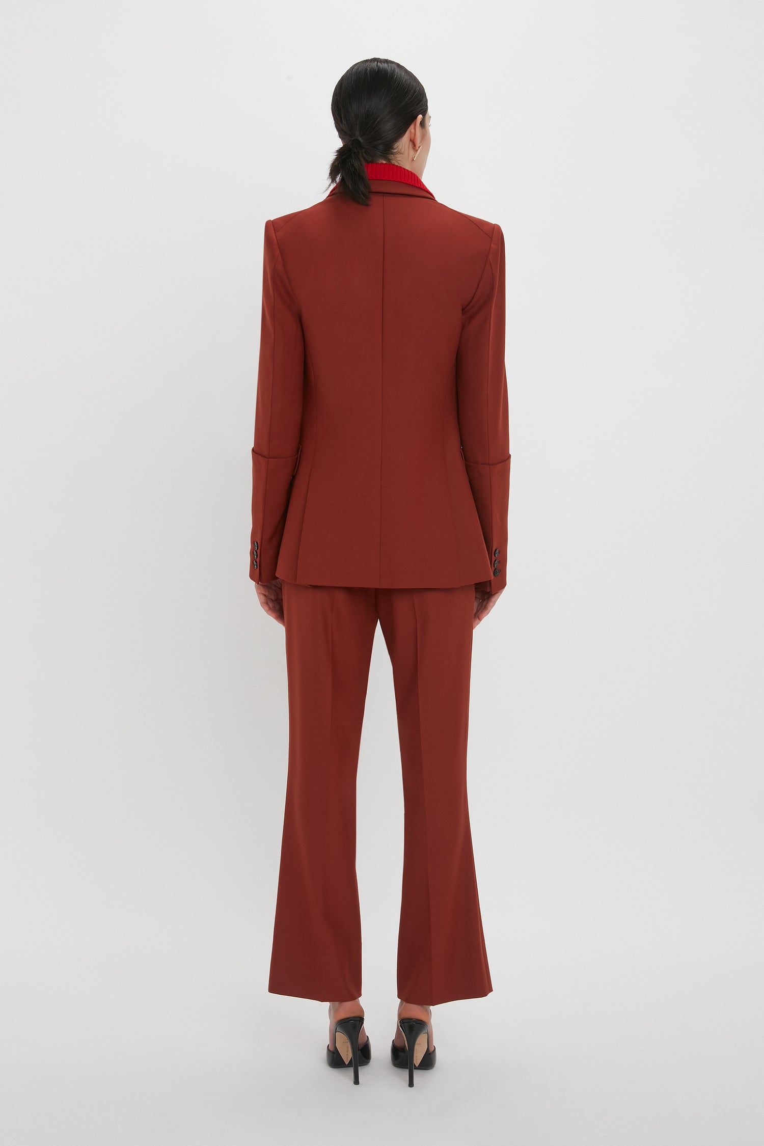A person with black hair in a ponytail is standing with their back to the camera, wearing a red tailored suit featuring Victoria Beckham's Wide Cropped Flare Trouser In Russet and black heeled shoes against a plain white background.