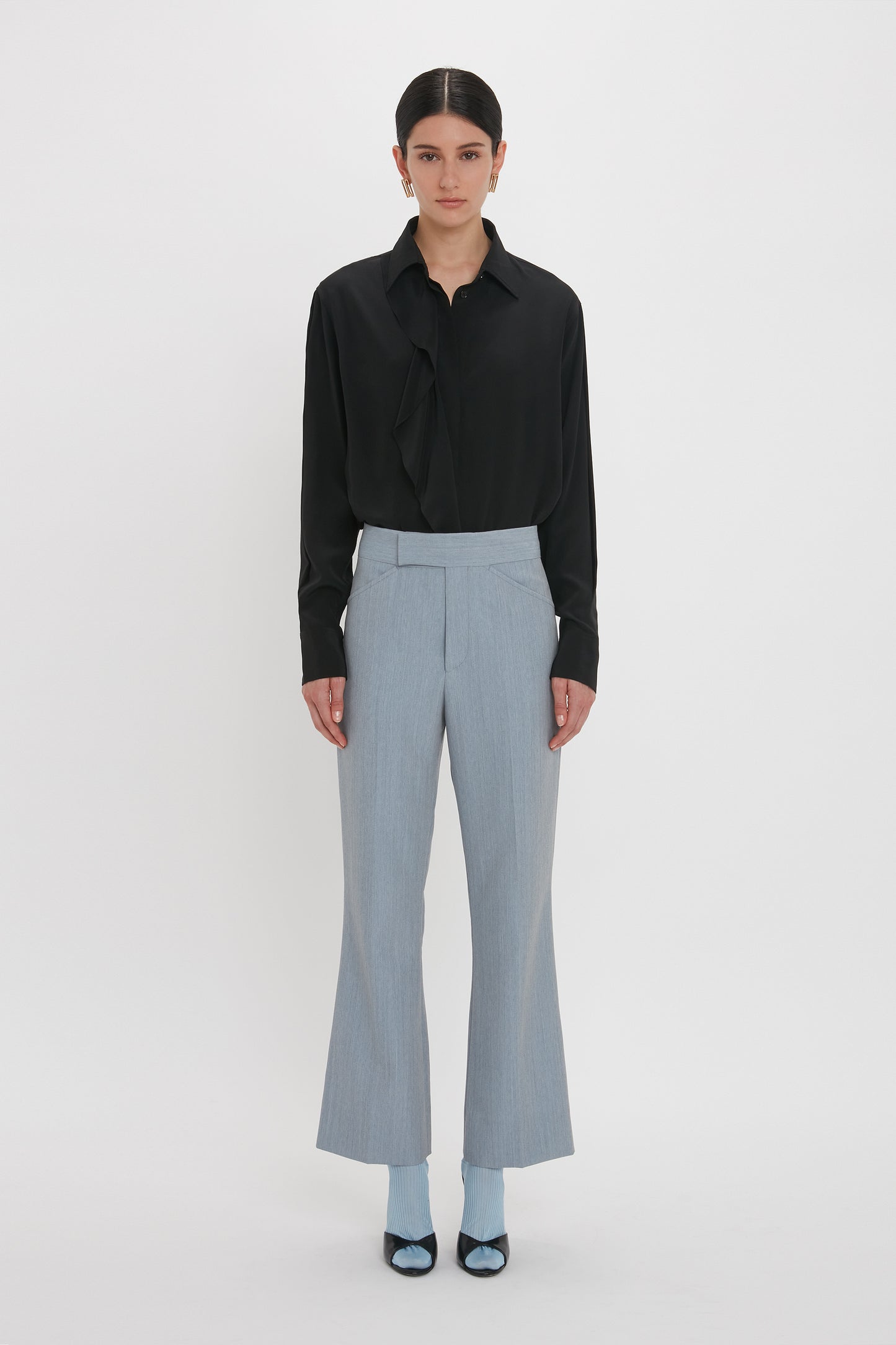 A person is standing against a plain white background, wearing a black button-up shirt, Victoria Beckham Exclusive Wide Cropped Flare Trouser In Marina in light gray, blue socks, and black shoes.