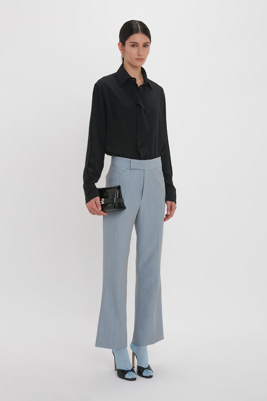 A woman stands in front of a plain white background, wearing a black blouse and Victoria Beckham Exclusive Wide Cropped Flare Trouser In Marina. She completes her outfit with matching blue open-toe heels and holds a black clutch bag.