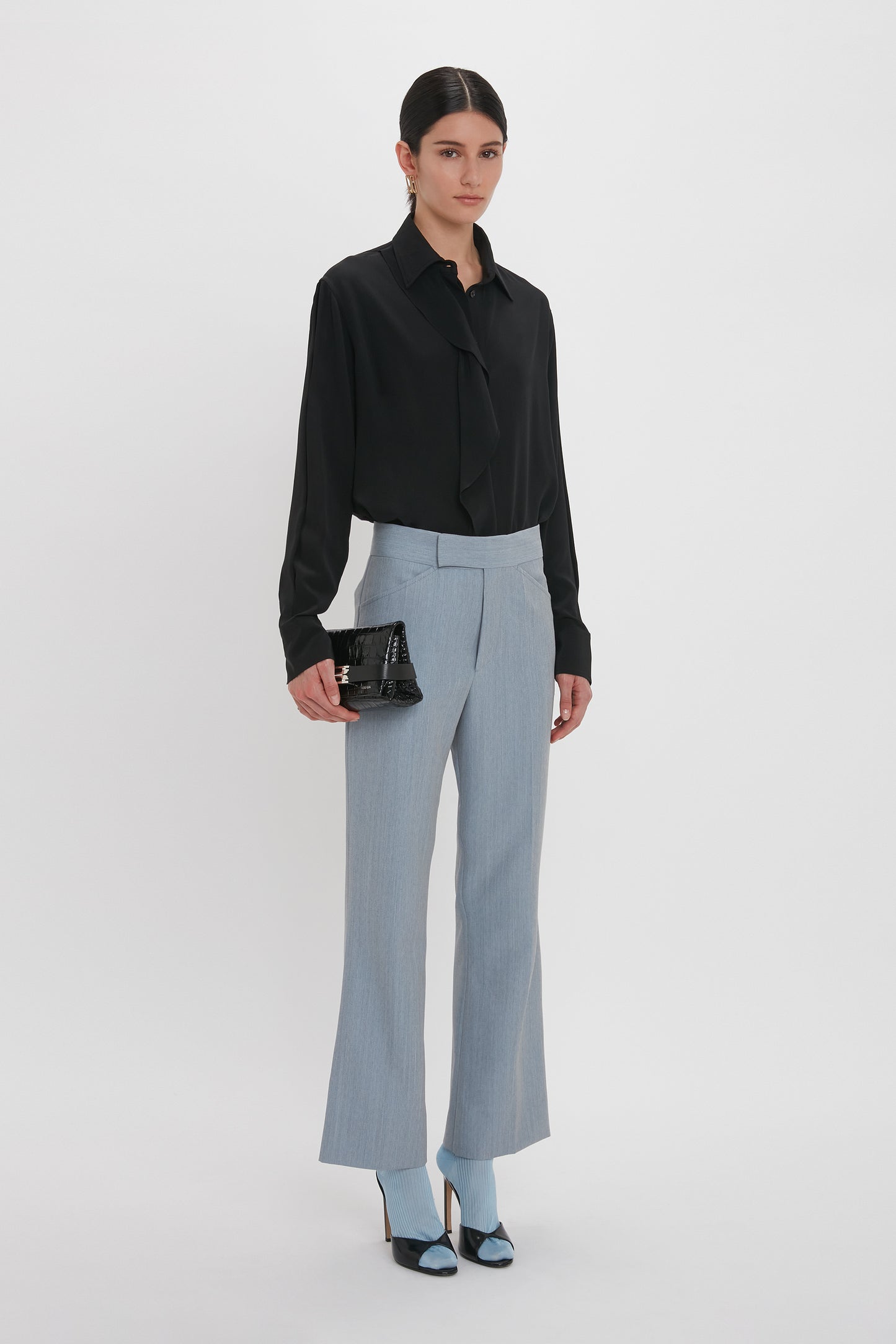 A person wearing a black blouse and light blue trousers, holding a Victoria Beckham Mini B Pouch Bag In Croc Effect Black Leather. They are standing against a plain white background.