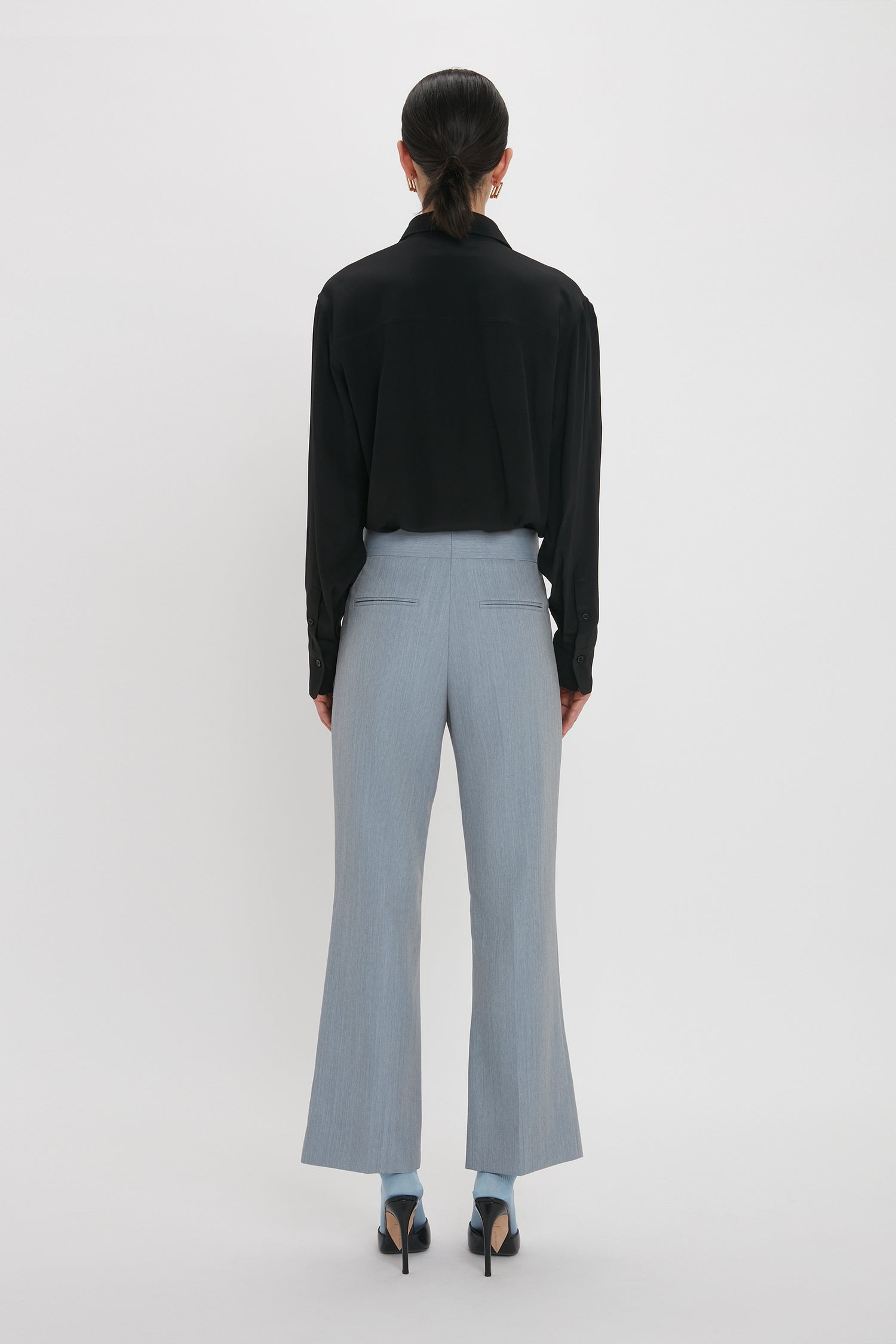 A person is standing facing away from the camera, wearing a black long-sleeve shirt, Victoria Beckham Exclusive Wide Cropped Flare Trouser In Marina, and black heels, with hair tied back.