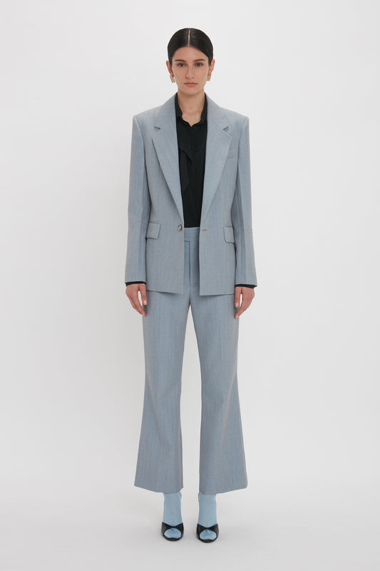 Person standing against a plain white background, impeccably tailored in a light gray Exclusive Sleeve Detail Patch Pocket Jacket In Marina by Victoria Beckham with contemporary detailing, featuring a black shirt and light blue socks paired with black and blue shoes.